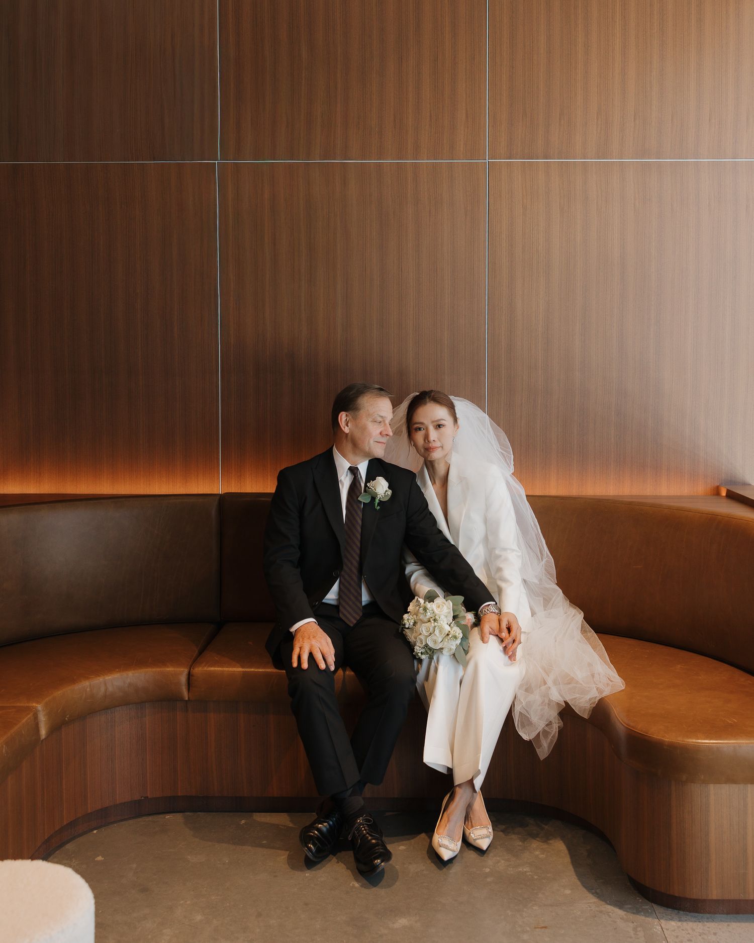 A bride in a white dress and veil sits with a groom in a black tuxedo on a curved leather bench in Nashville Tennesee.