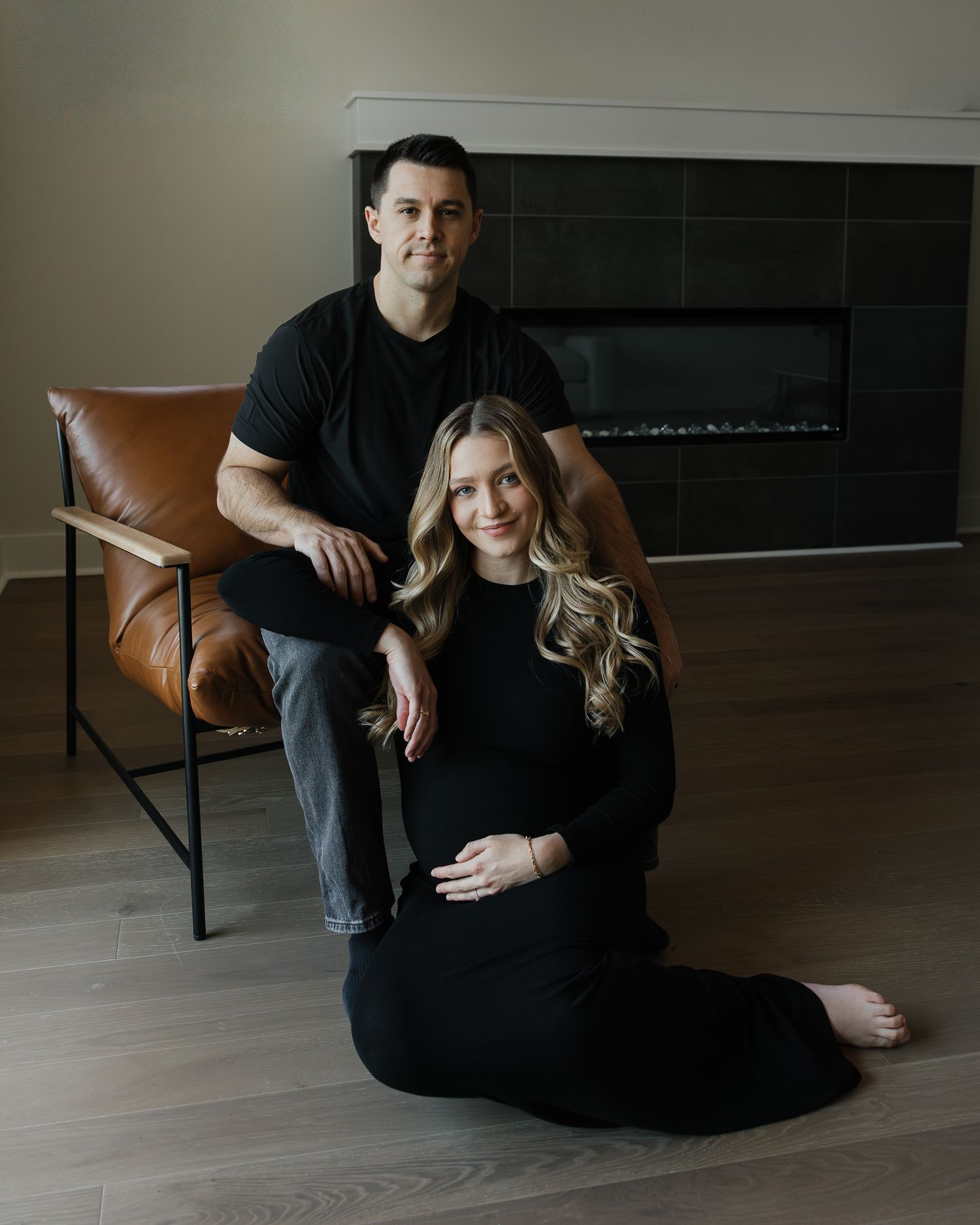 Couple in black outfits sitting together in a minimalist dark room with moody lighting.