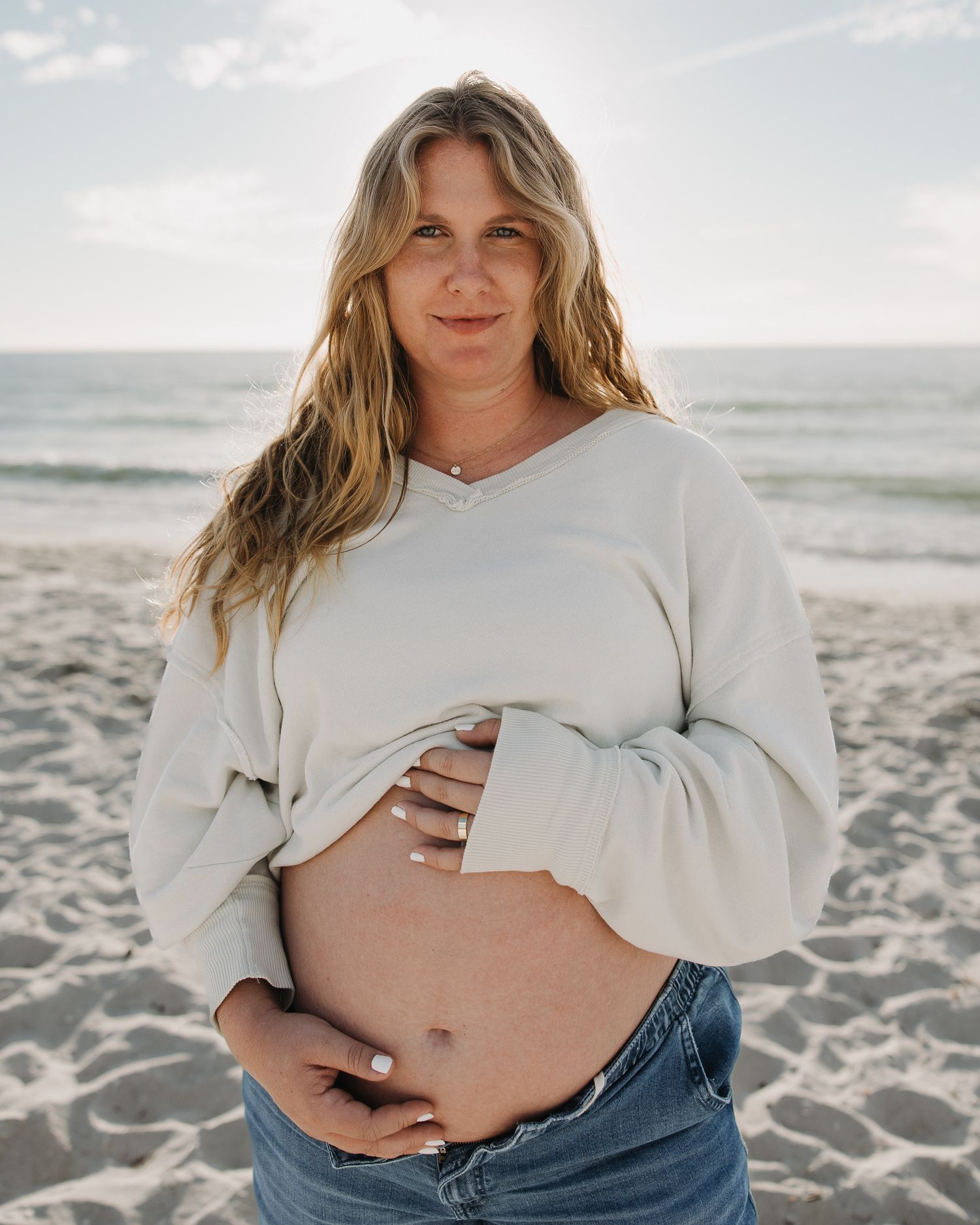 Expectant parent in white sweater and jeans poses on sunny beach during maternity photoshoot.