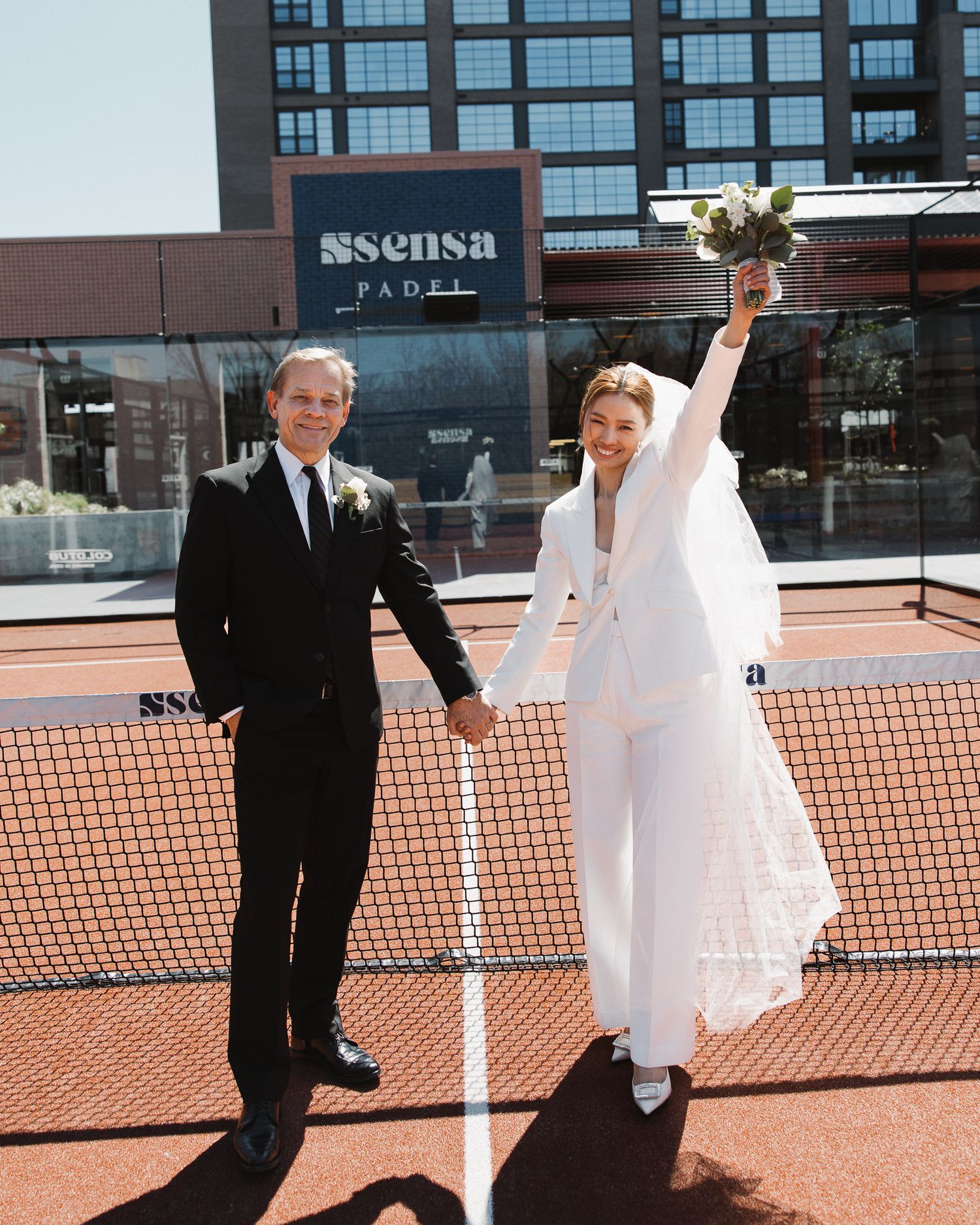 Bride in white pantsuit and groom in black tuxedo holding hands on a tennis court, celebrating their wedding day in Downtown Nashville.