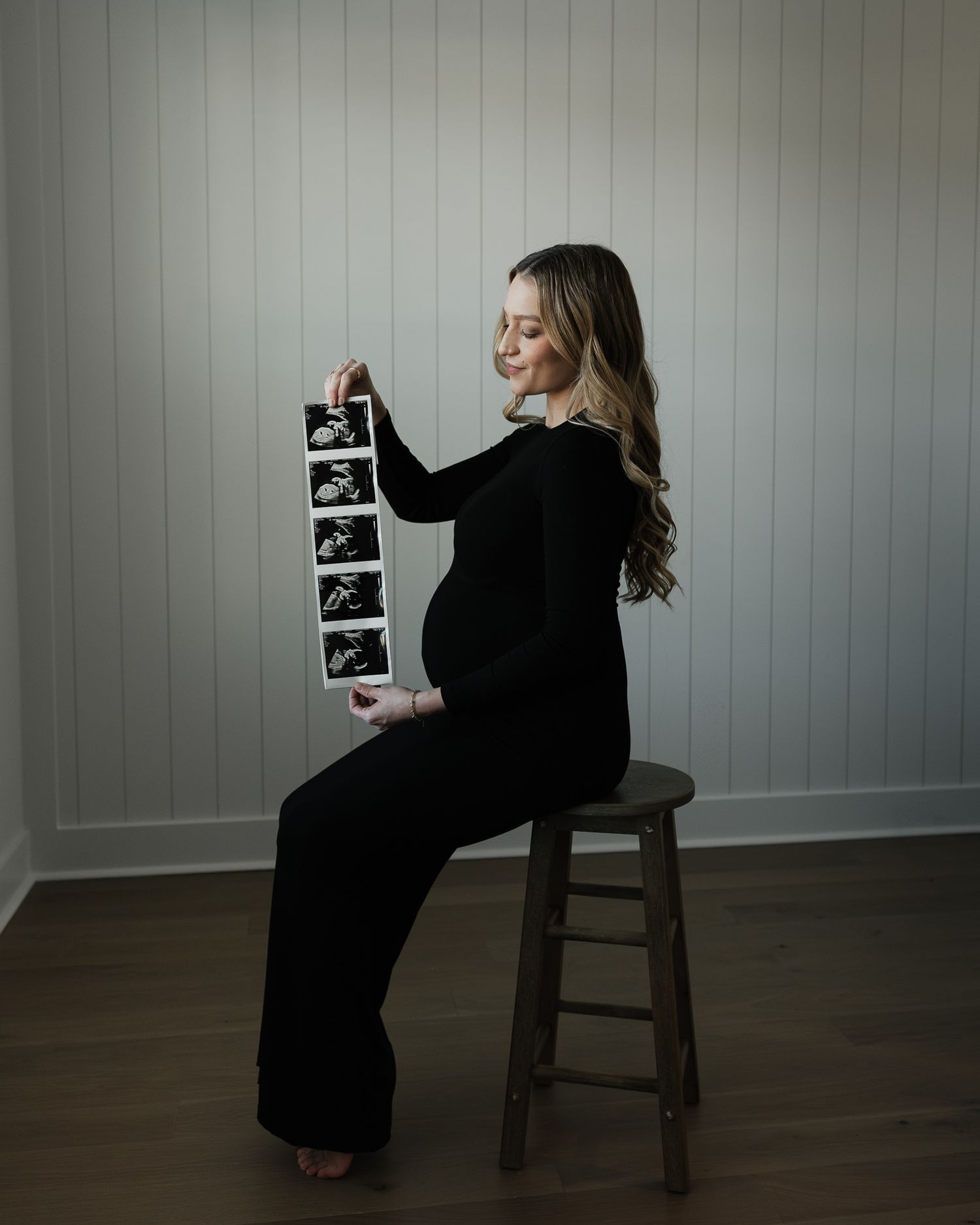 Expectant mother in black dress sits on wooden stool holding ultrasound photos against white paneled wall.