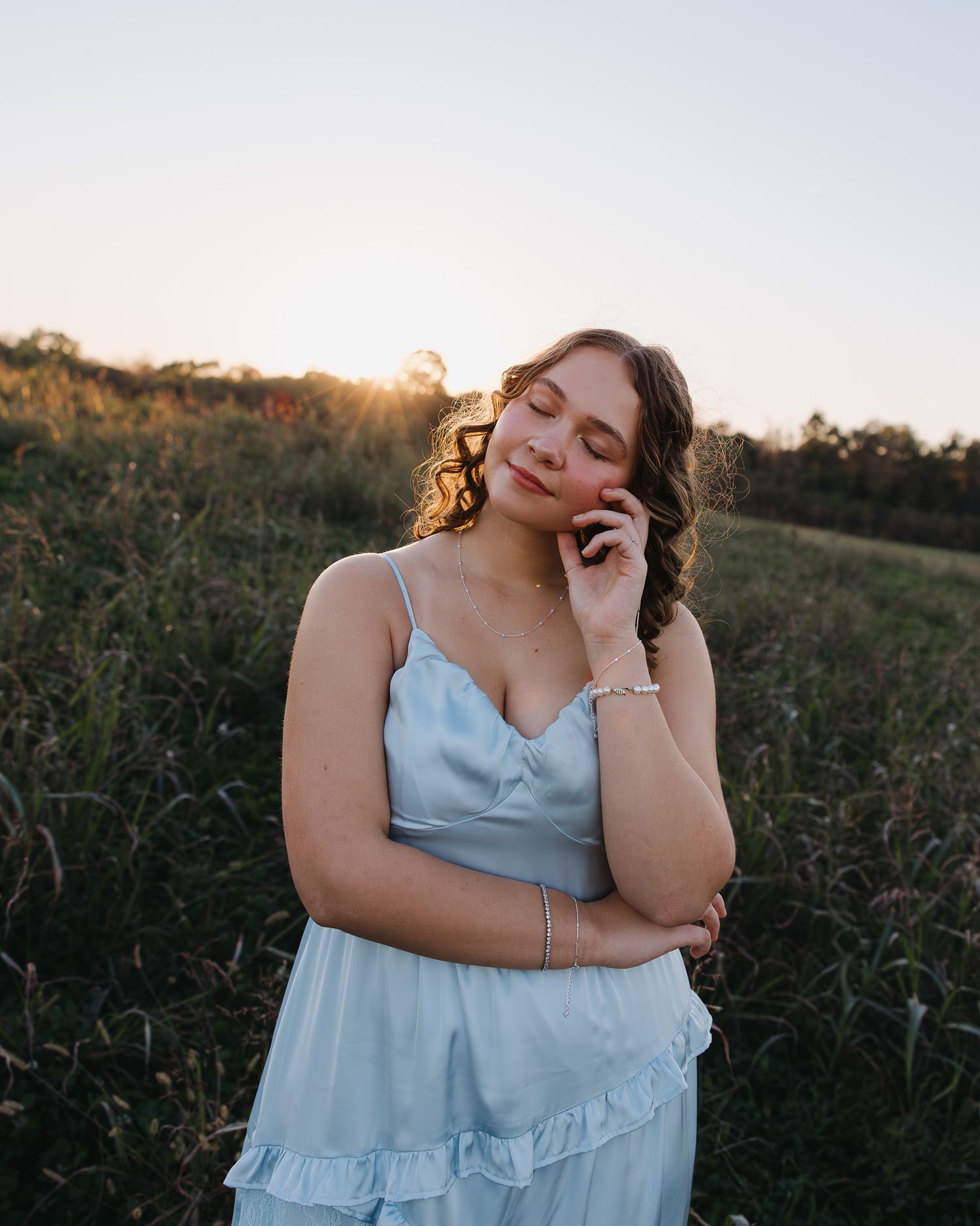 A woman in a light blue dress poses playfully in a field at sunset, creating a dreamy outdoor portrait series.