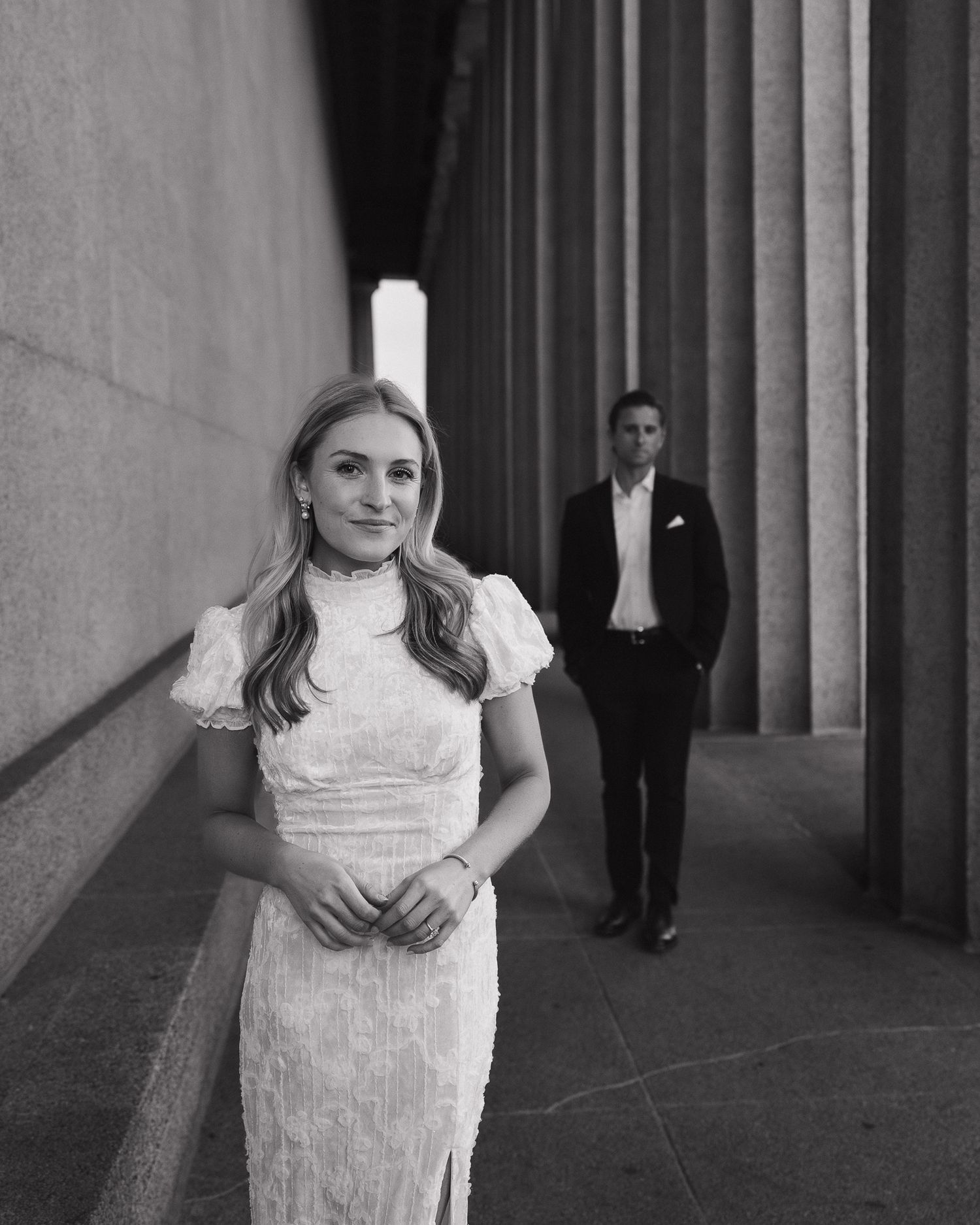 A black and white engagement photo showing a woman in a white dress and man standing in a columned corridor.