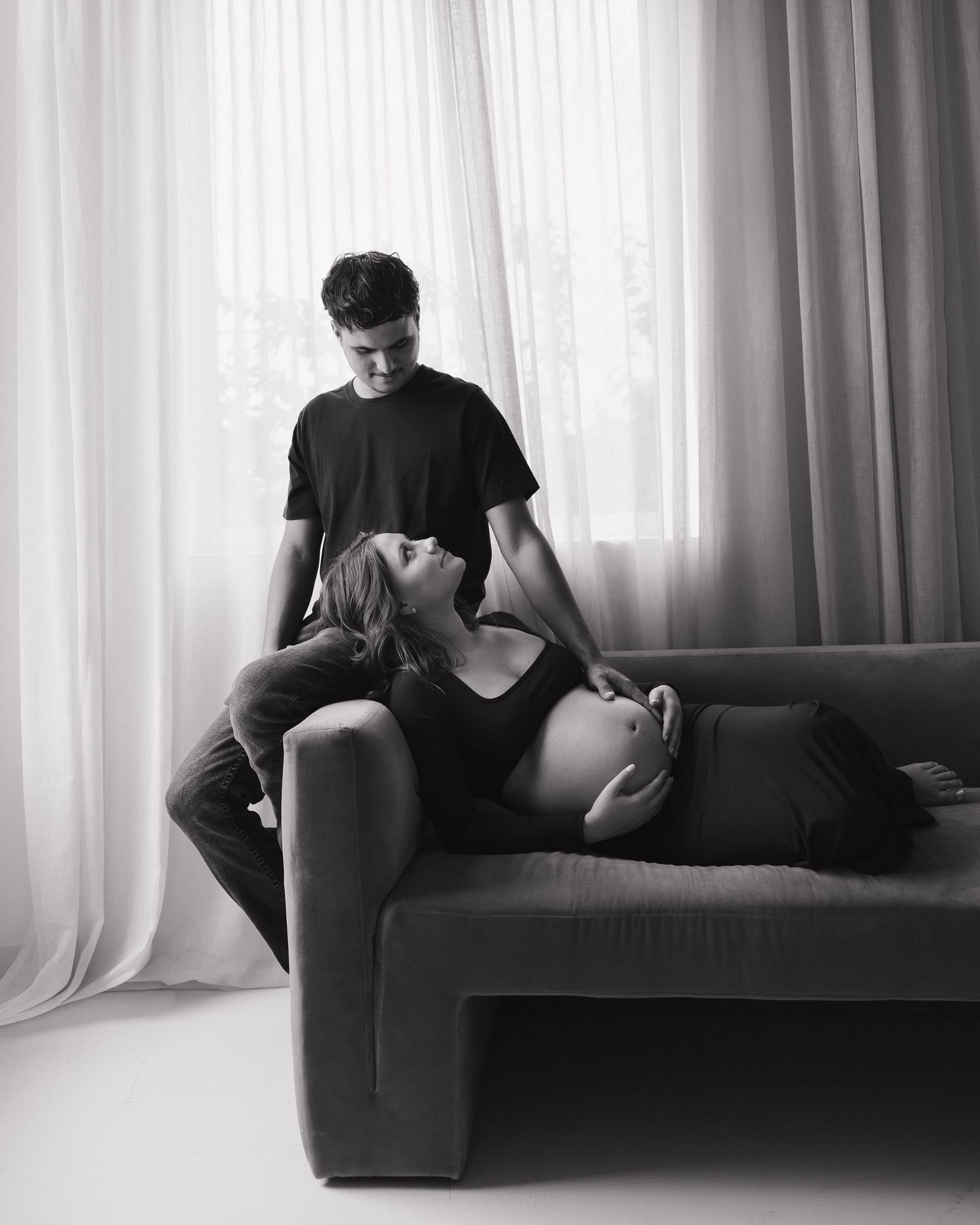 Black and white maternity photo of pregnant woman lying on sofa with partner gazing down at her bump.