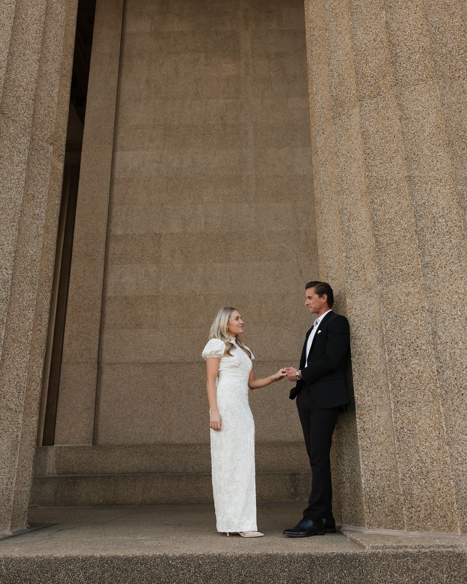 A man and woman share a candid moment between stone pillars in an architectural setting.
