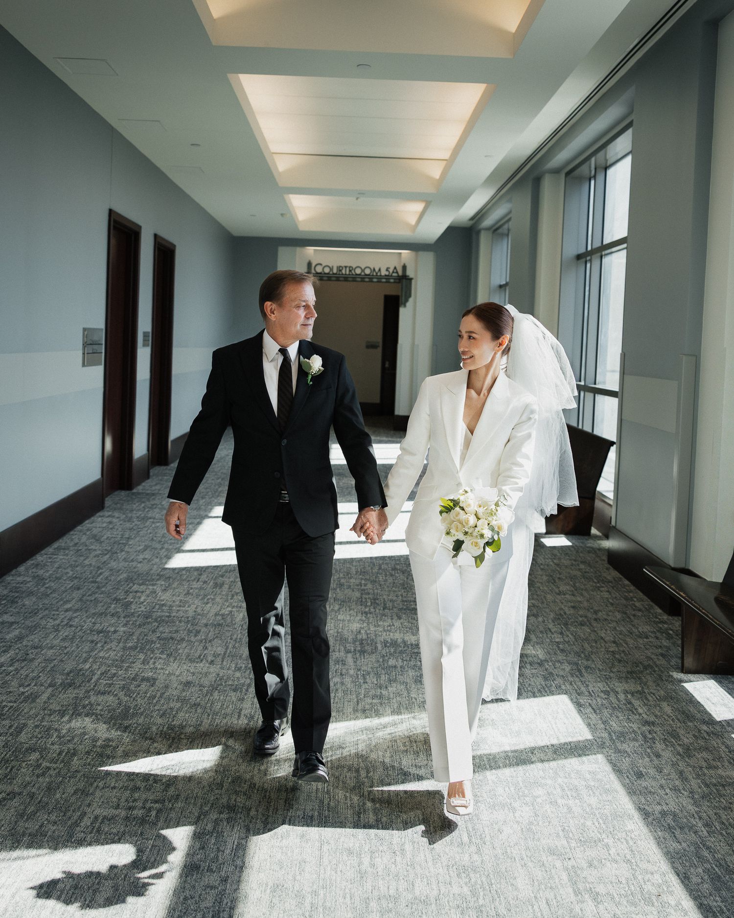 Bride in white pantsuit holding bouquet walks with groom in black tuxedo through sunlit courthouse hallway in Nashville Tennessee.