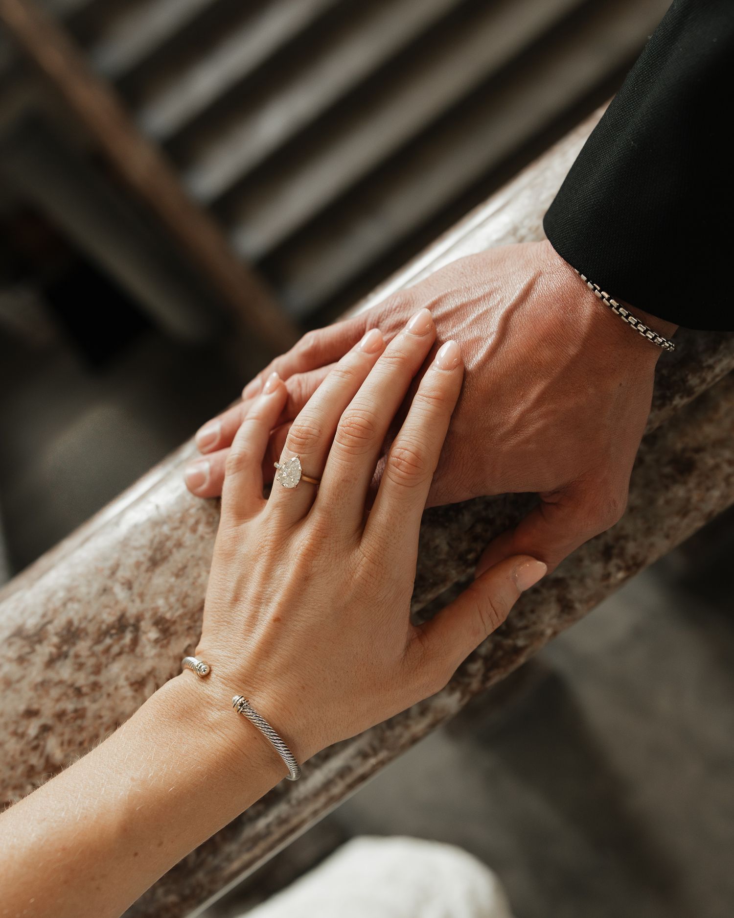 Two hands touch tenderly on a stone railing with one hand wearing an engagement ring and delicate bracelet.