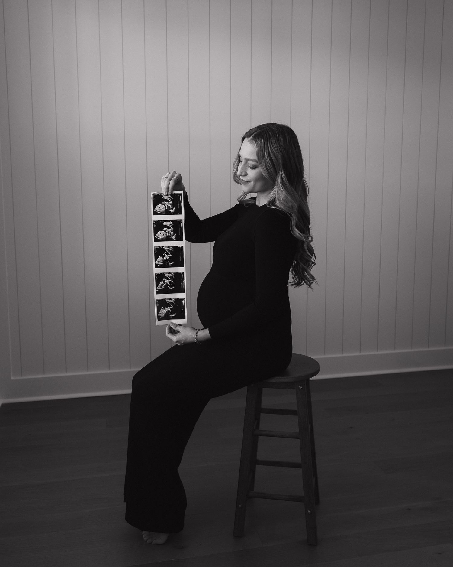 A black and white maternity photo showing a pregnant lady in dark clothing posing with a chair against a striped wall.