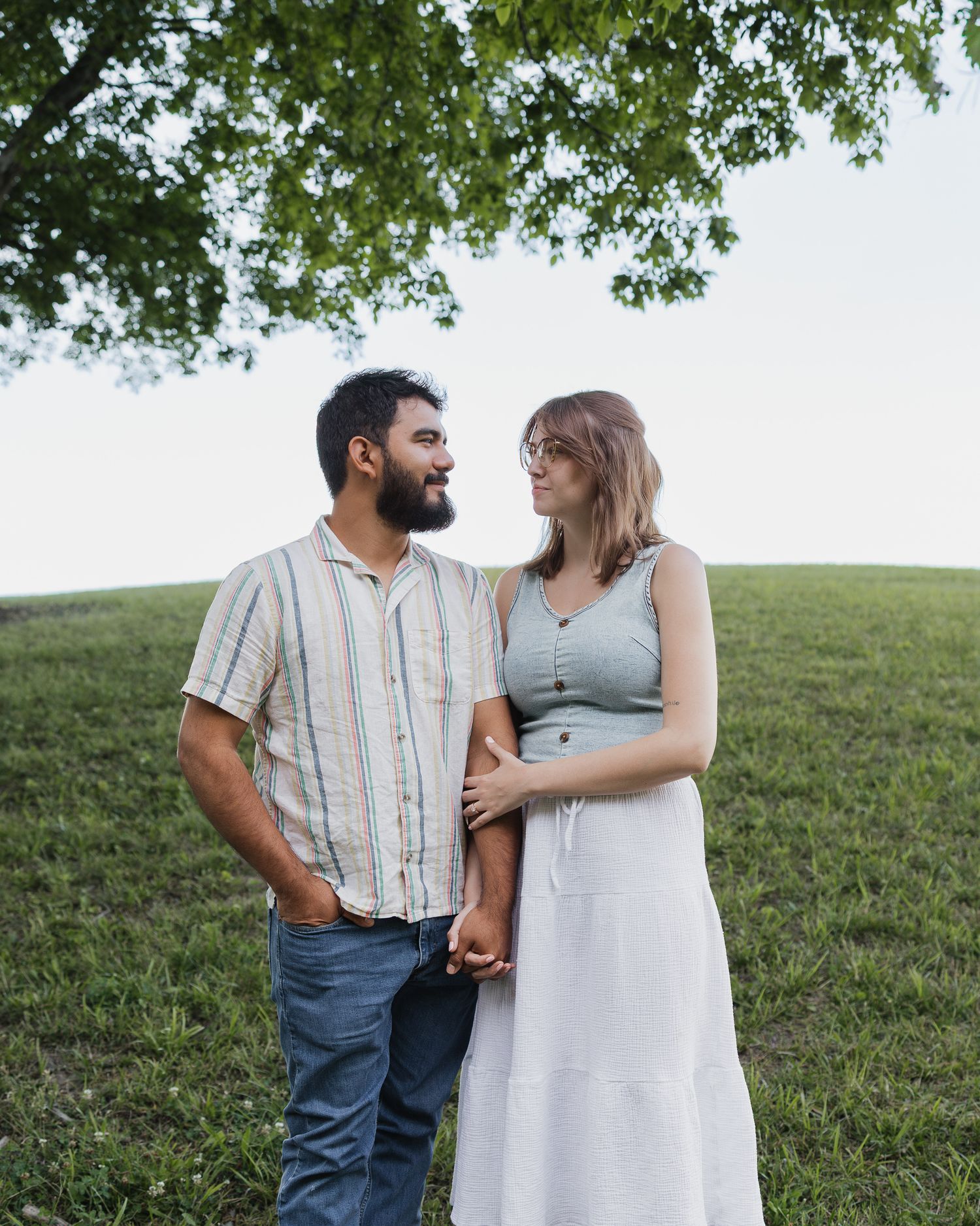 A couple stands on a green hillside under a tree, holding hands and gazing at each other affectionately.