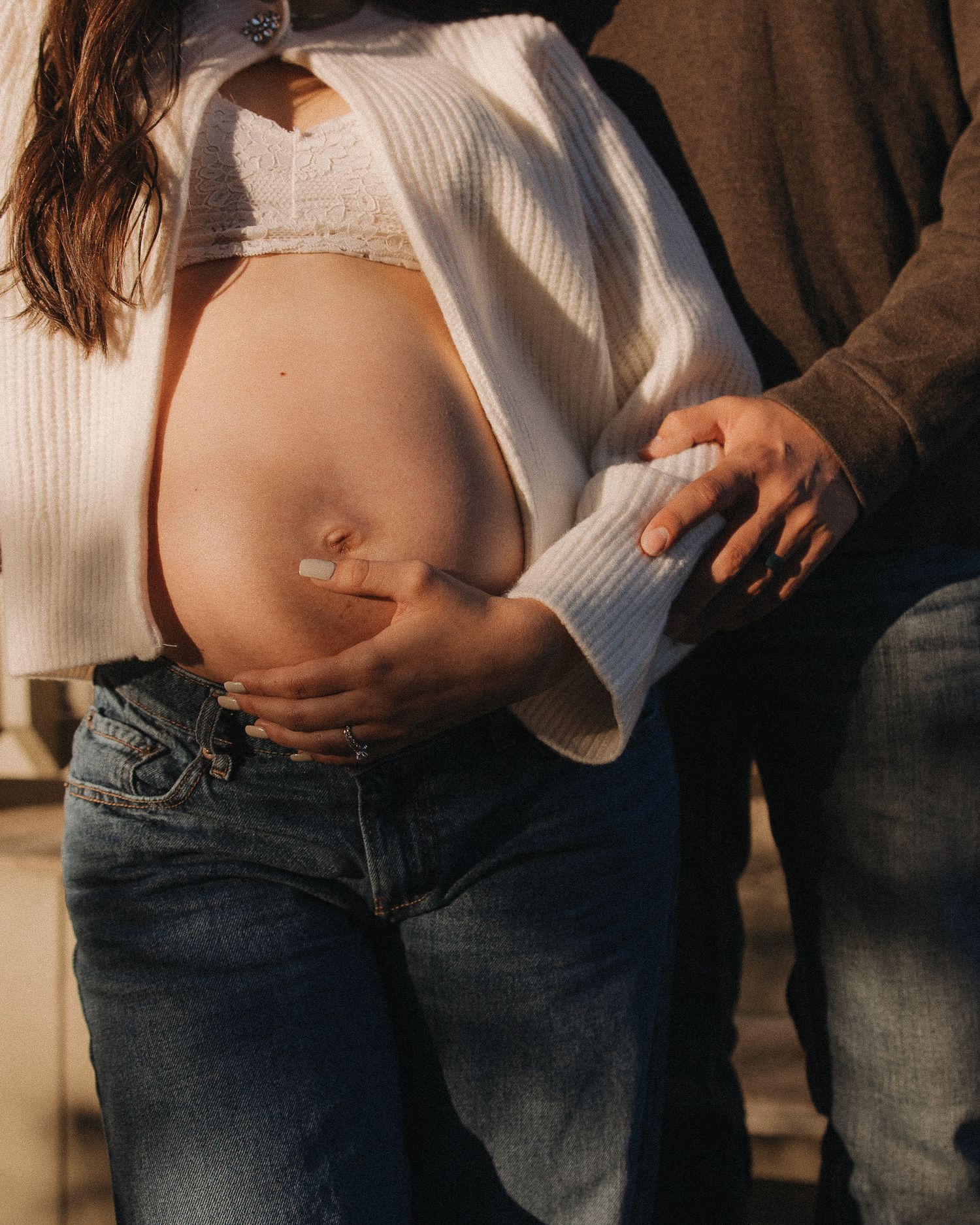 Intimate maternity photoshoot showing a couple in casual attire with white sweater and jeans against a warm background.