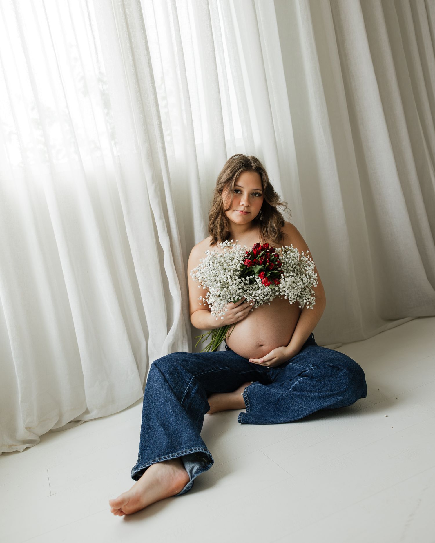 Pregnant woman sitting on floor holding white and red flowers against her bare belly, wearing blue jeans.