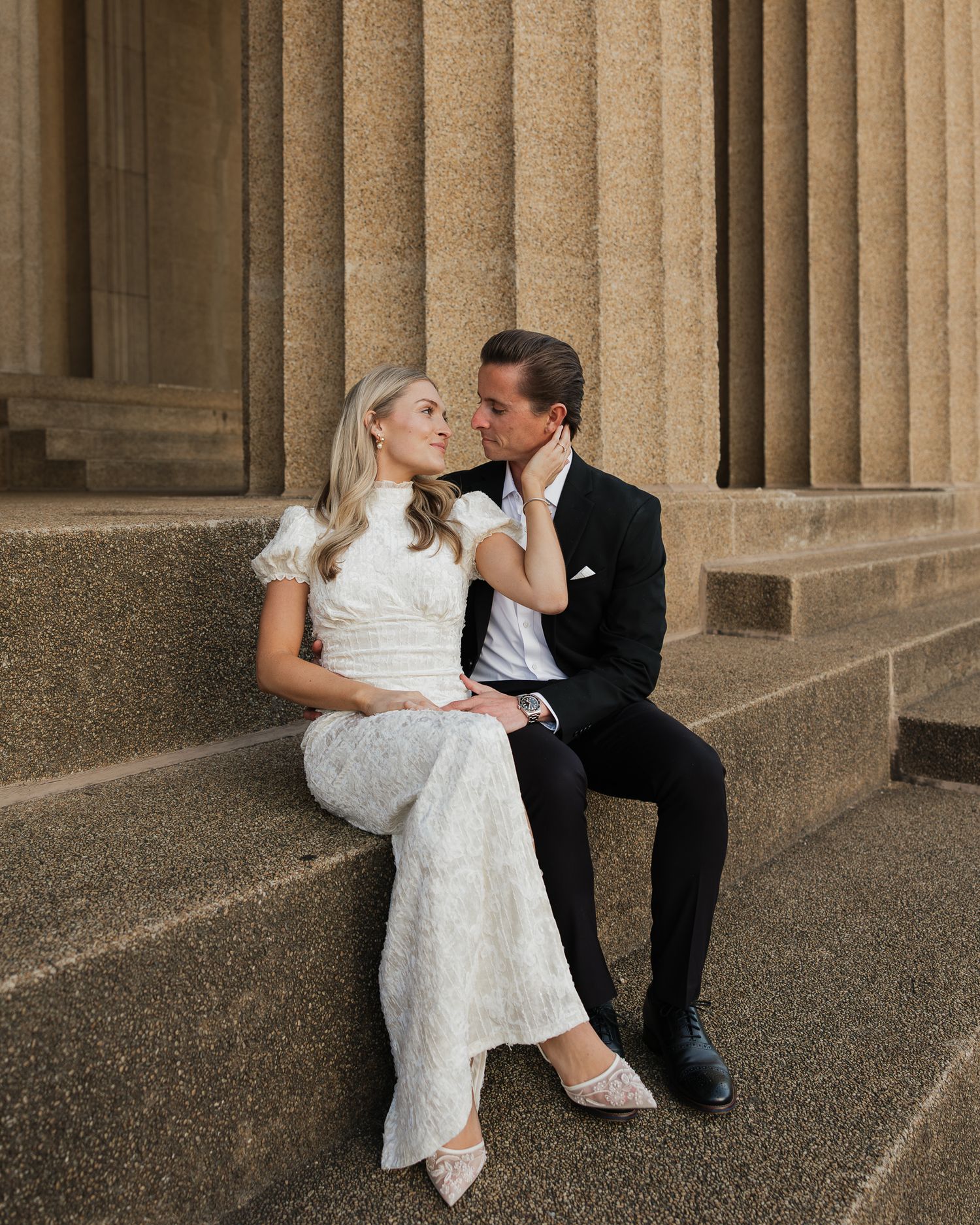Man and woman share intimate kisses while sitting on stone steps with classical columns in the background.