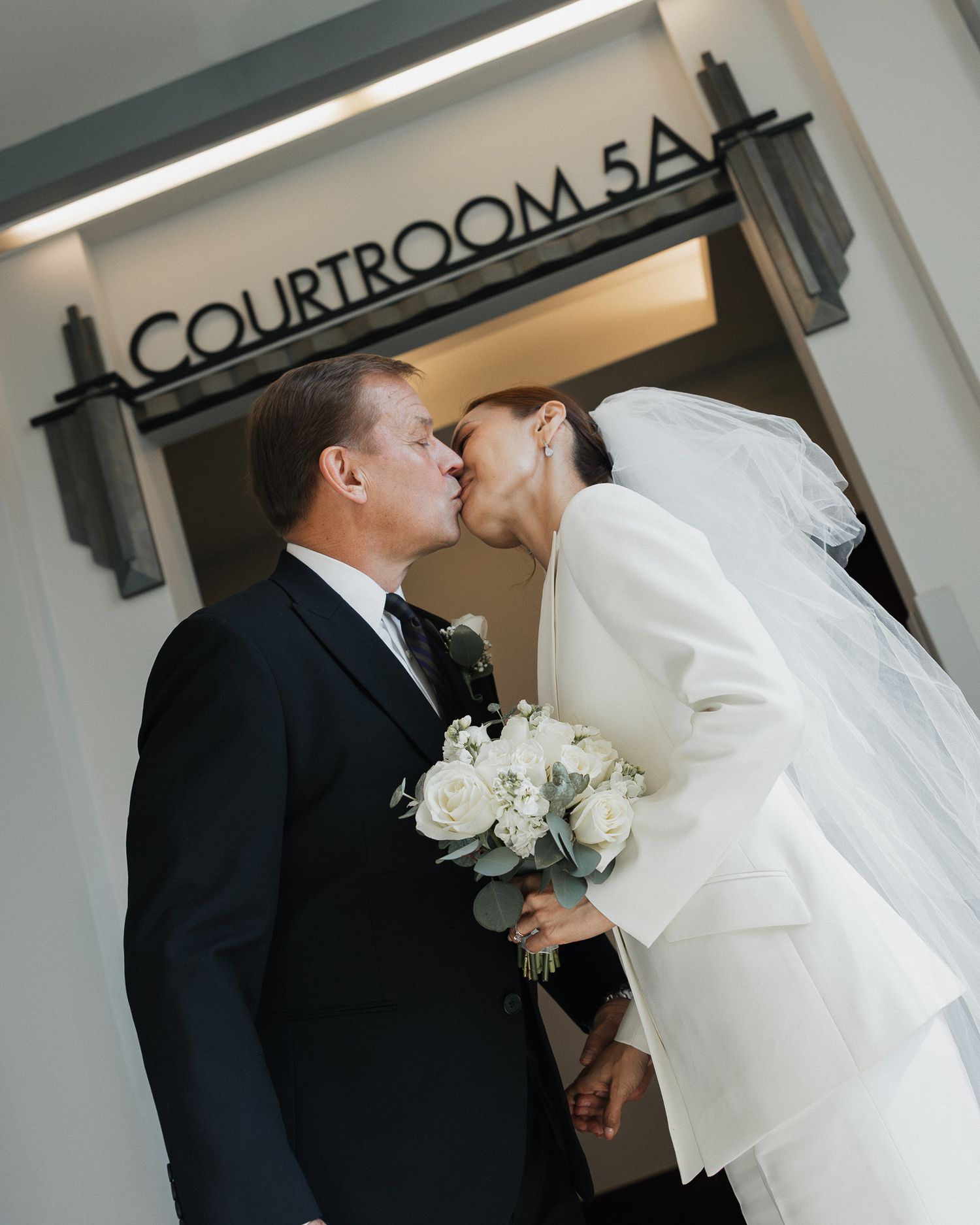 Bride and groom share a kiss outside Courtroom 5A, bride holding white roses bouquet wearing veil and white coat.
