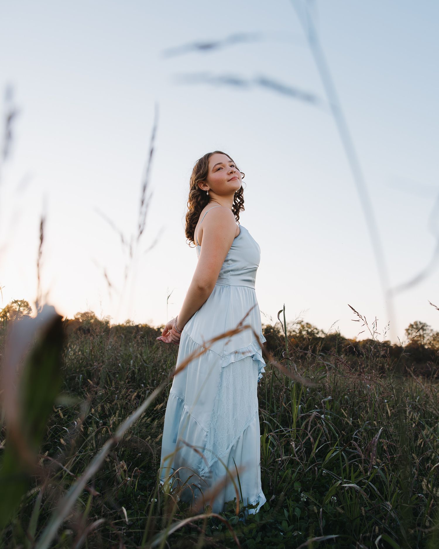 A high school senior in flowing light blue dress twirls in a field at sunset.