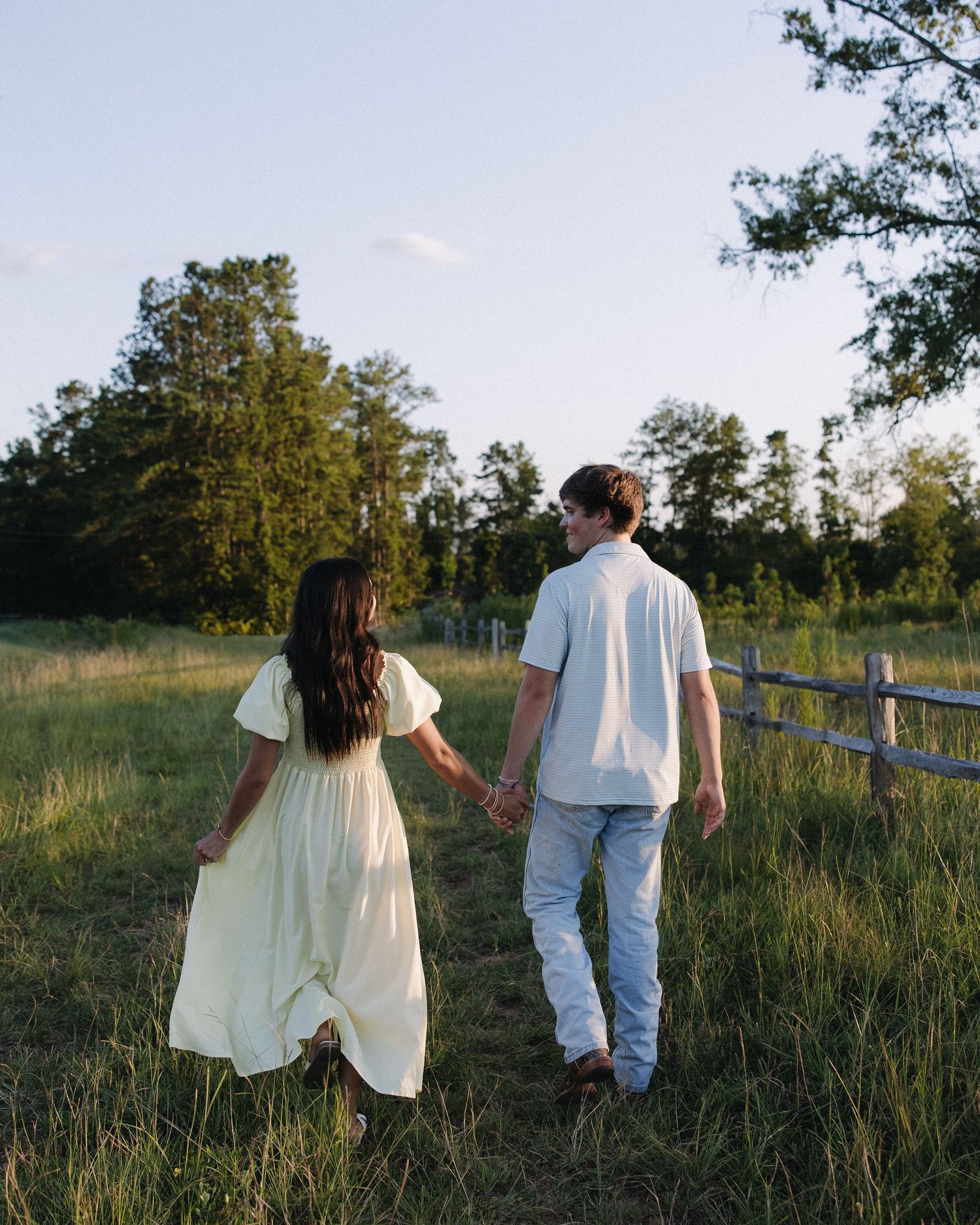 Couple walking hand in hand through a rustic meadow with wooden fence at sunset.