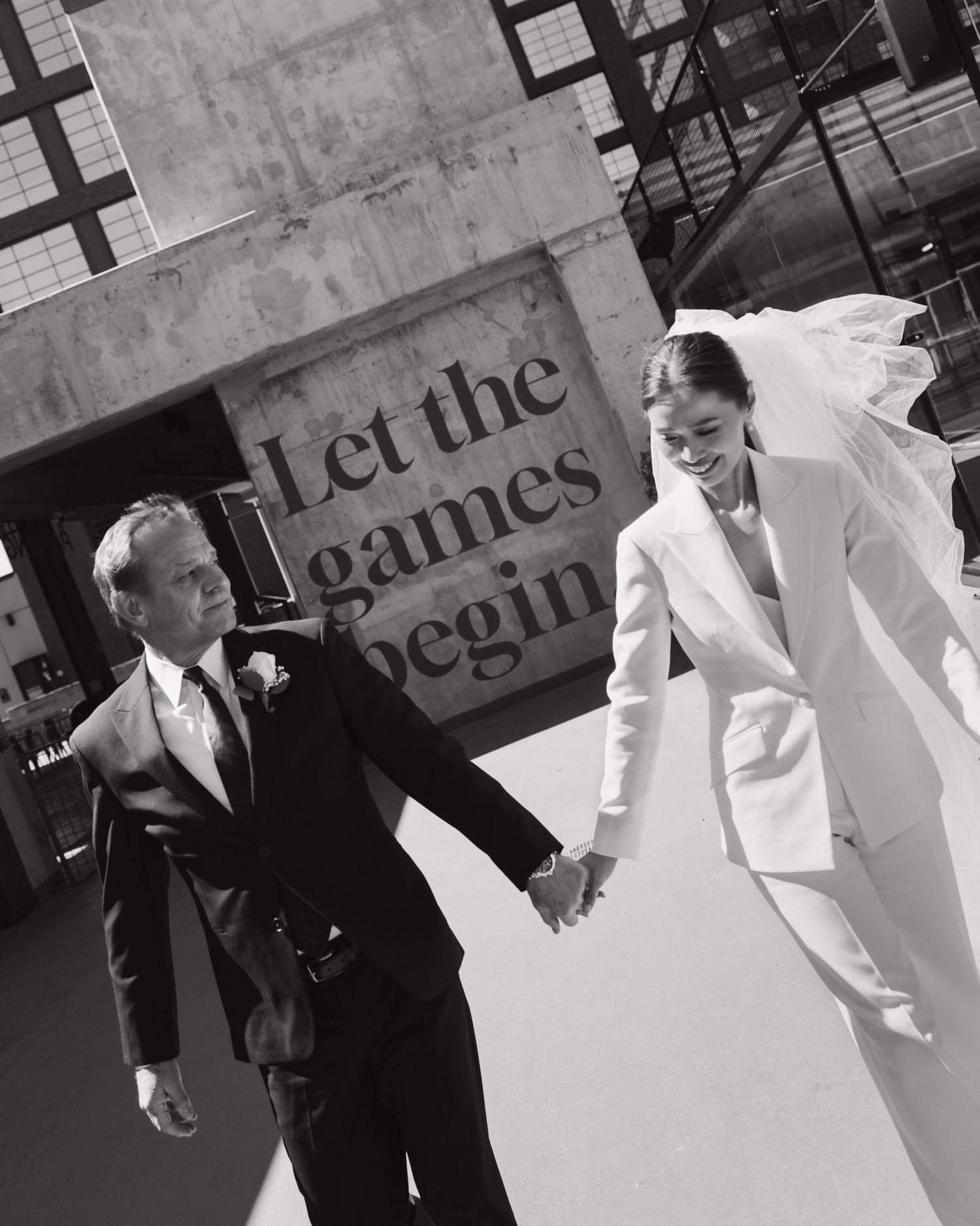 Black and white wedding photo of bride in white suit and holding hands with groom near 'Let the games begin' sign in Nashville Tennesee.