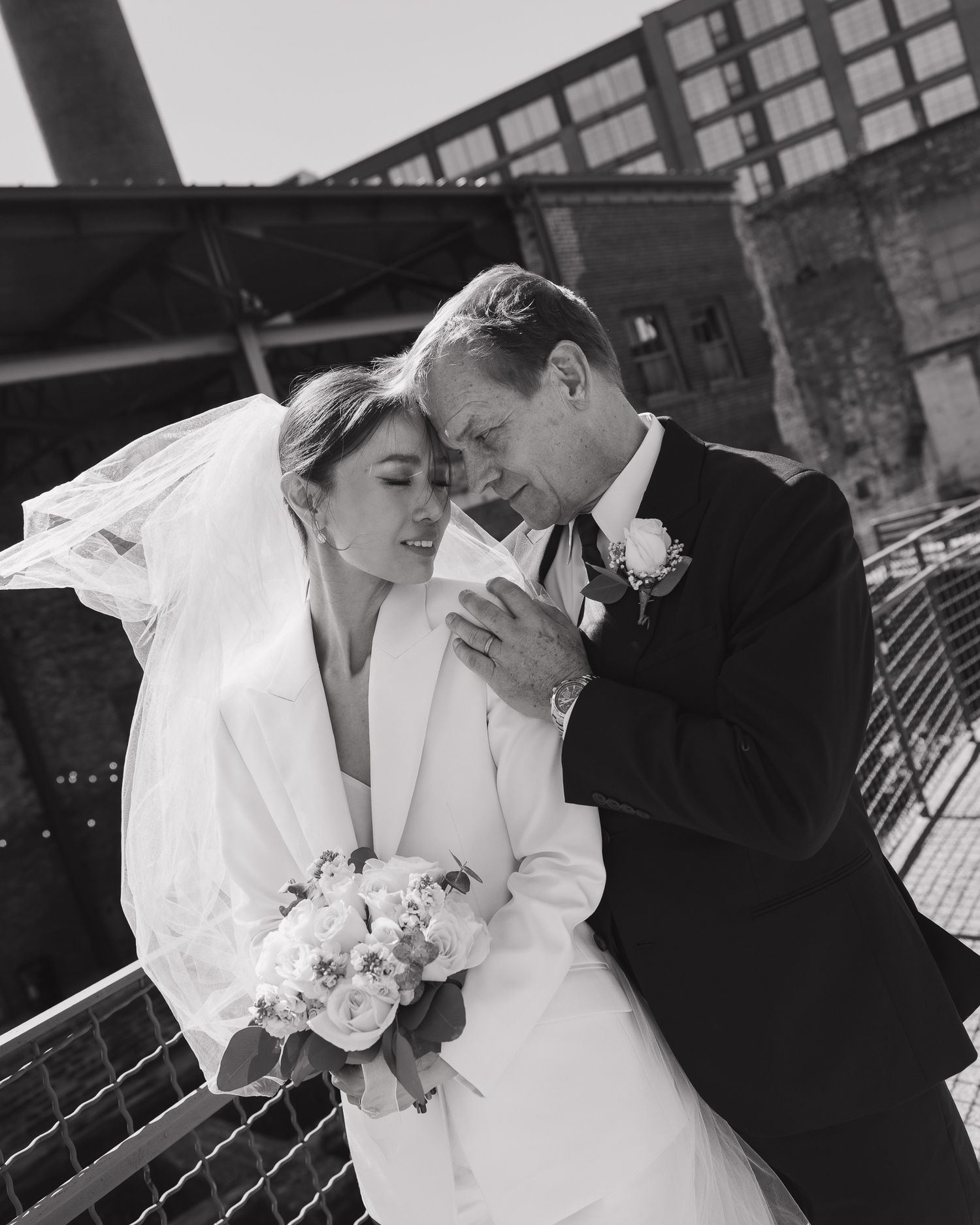 Black and white wedding photo of bride in white suit and groom in tuxedo sharing an intimate moment outdoors.