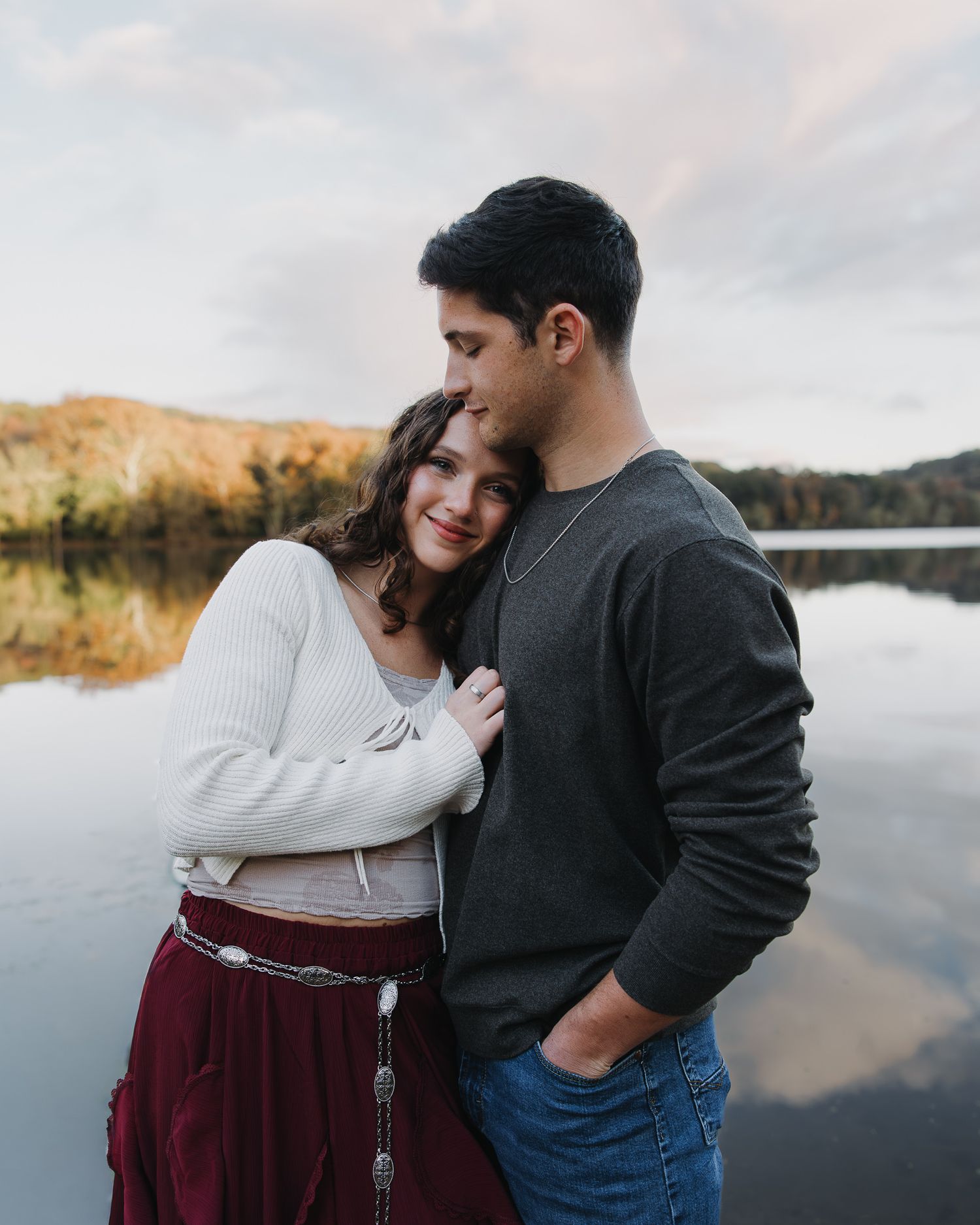 Couple embracing by a serene lake during golden hour with autumn colors in the background.