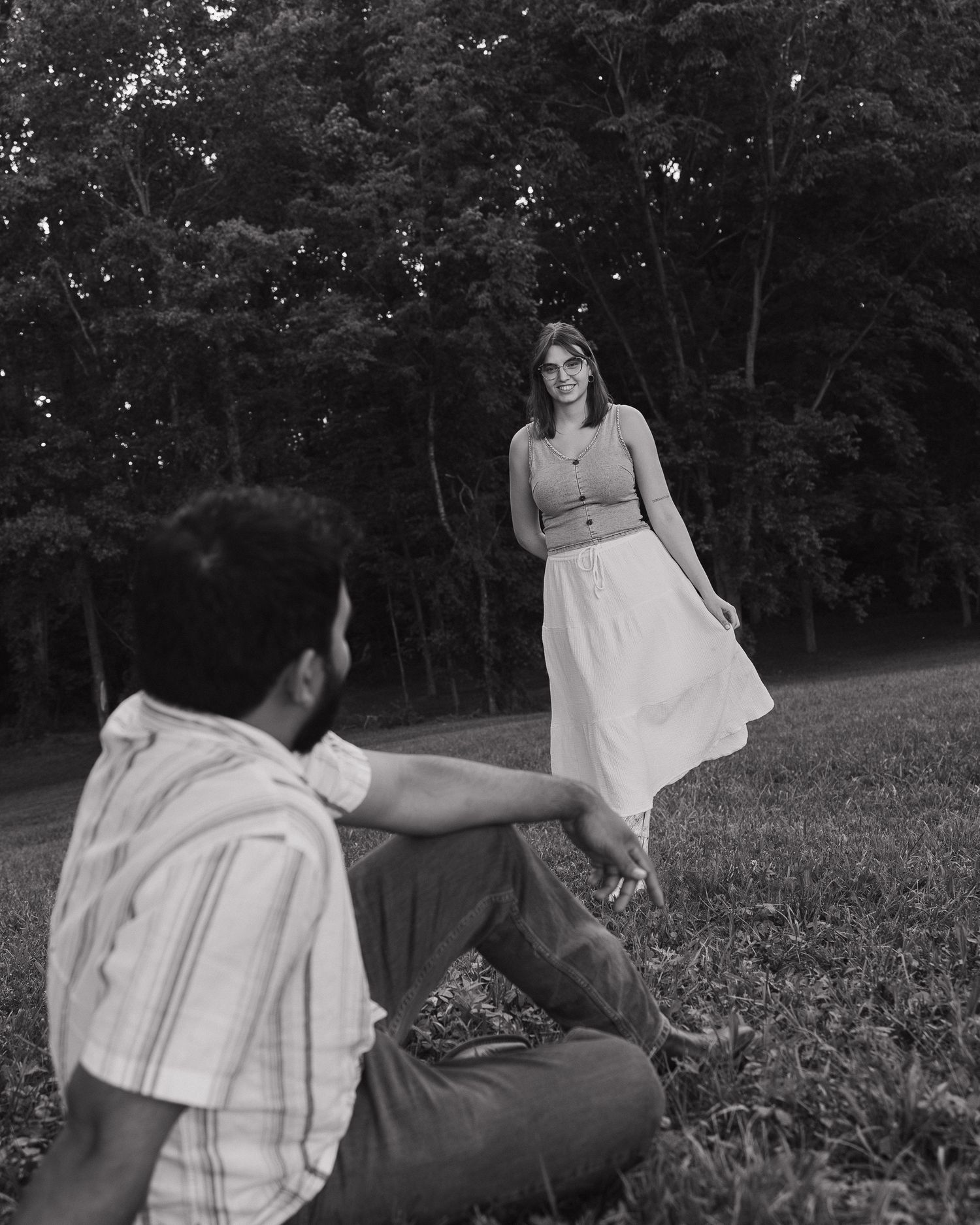 Black and white photo of a couple in a park, woman in skirt standing while man sits on grass.