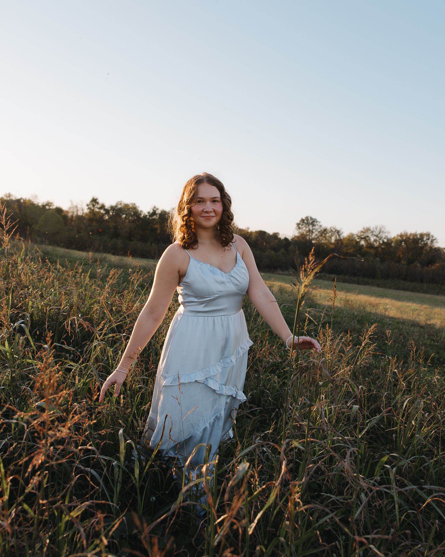 Person in flowing blue dress stands among tall grass at sunset.