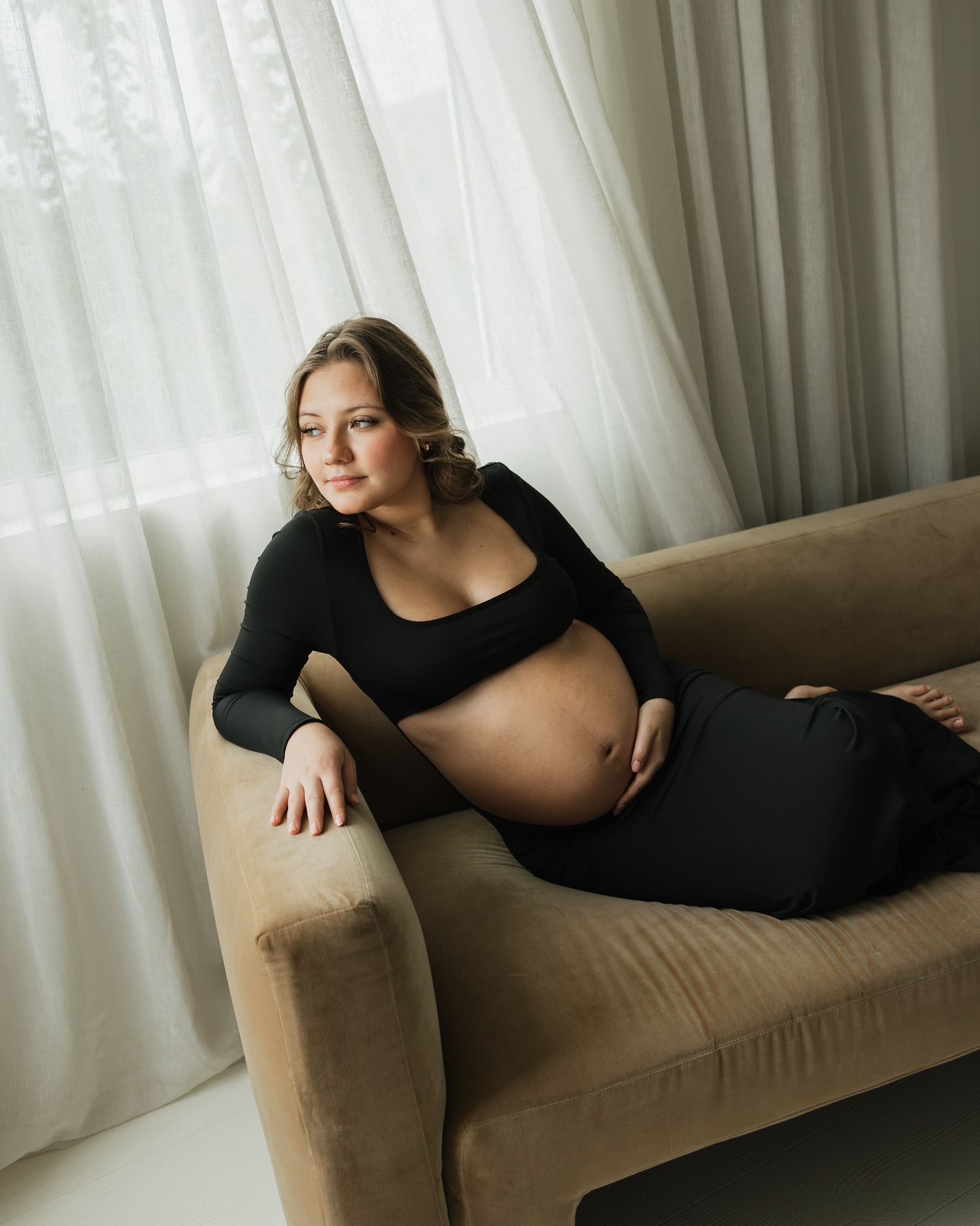 Pregnant woman in black dress reclines elegantly on a beige velvet sofa near sheer white curtains.
