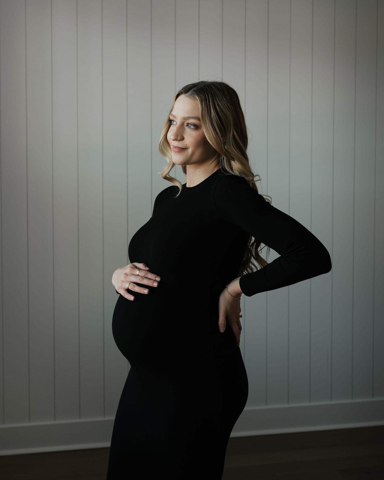 A pregnant woman in a black dress poses against a gray wall in a moody maternity portrait.