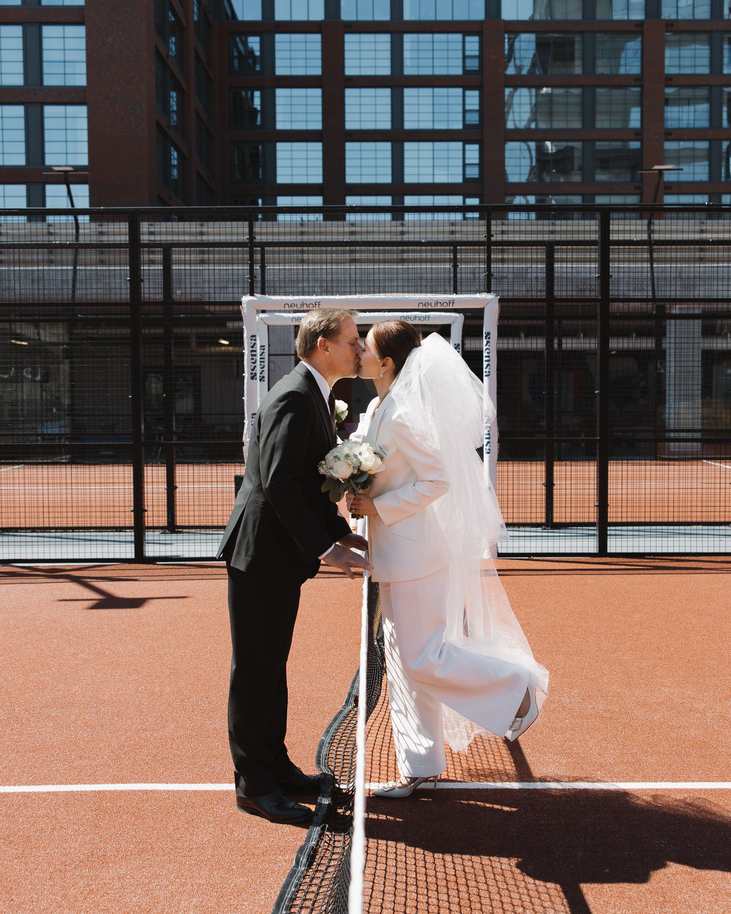 Bride and groom kissing at a tennis net on an outdoor court, with a building in the background in Nashville Tennessee.