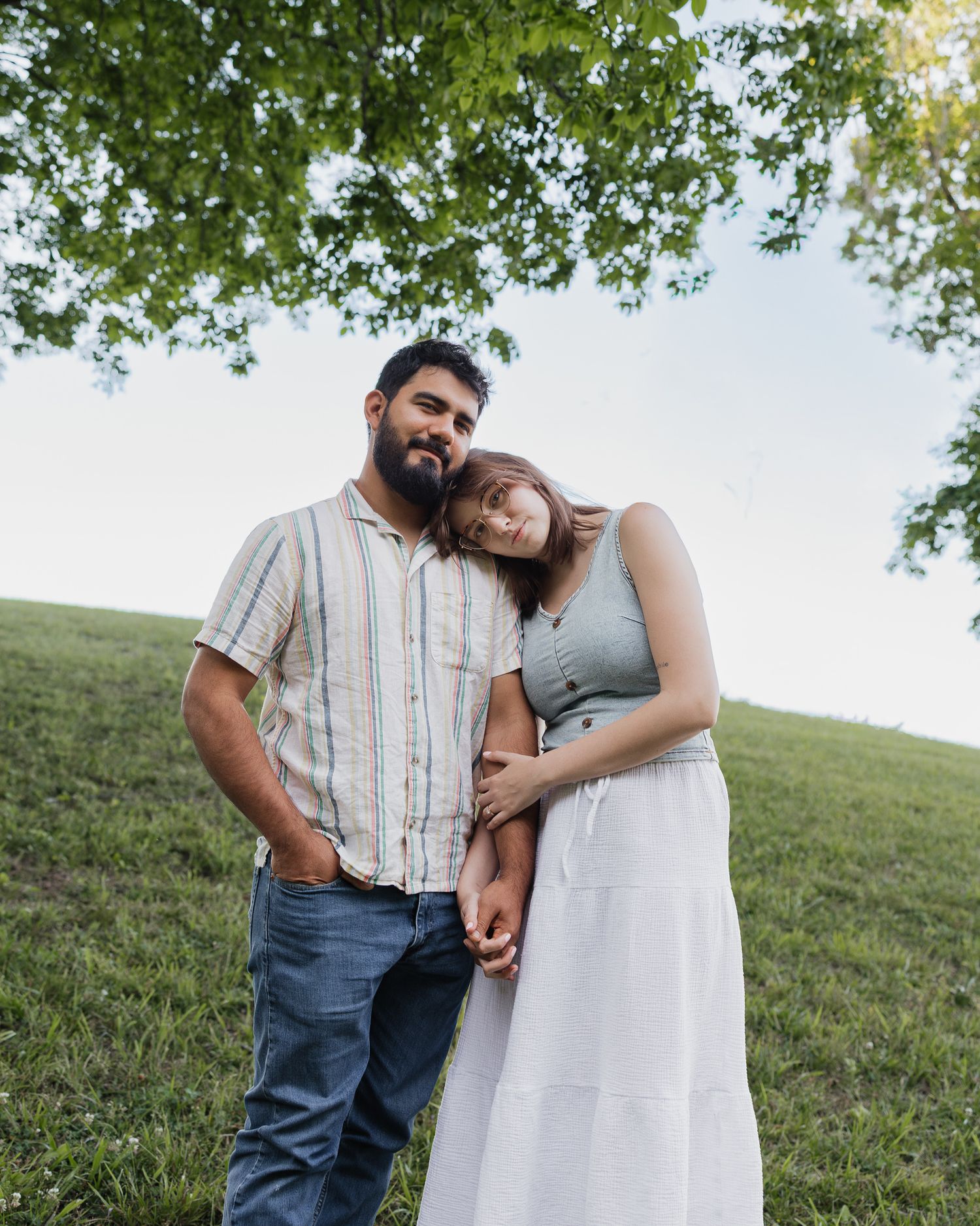 A couple stands embracing on a green hillside under a tree, the woman resting her head on the man's shoulder.