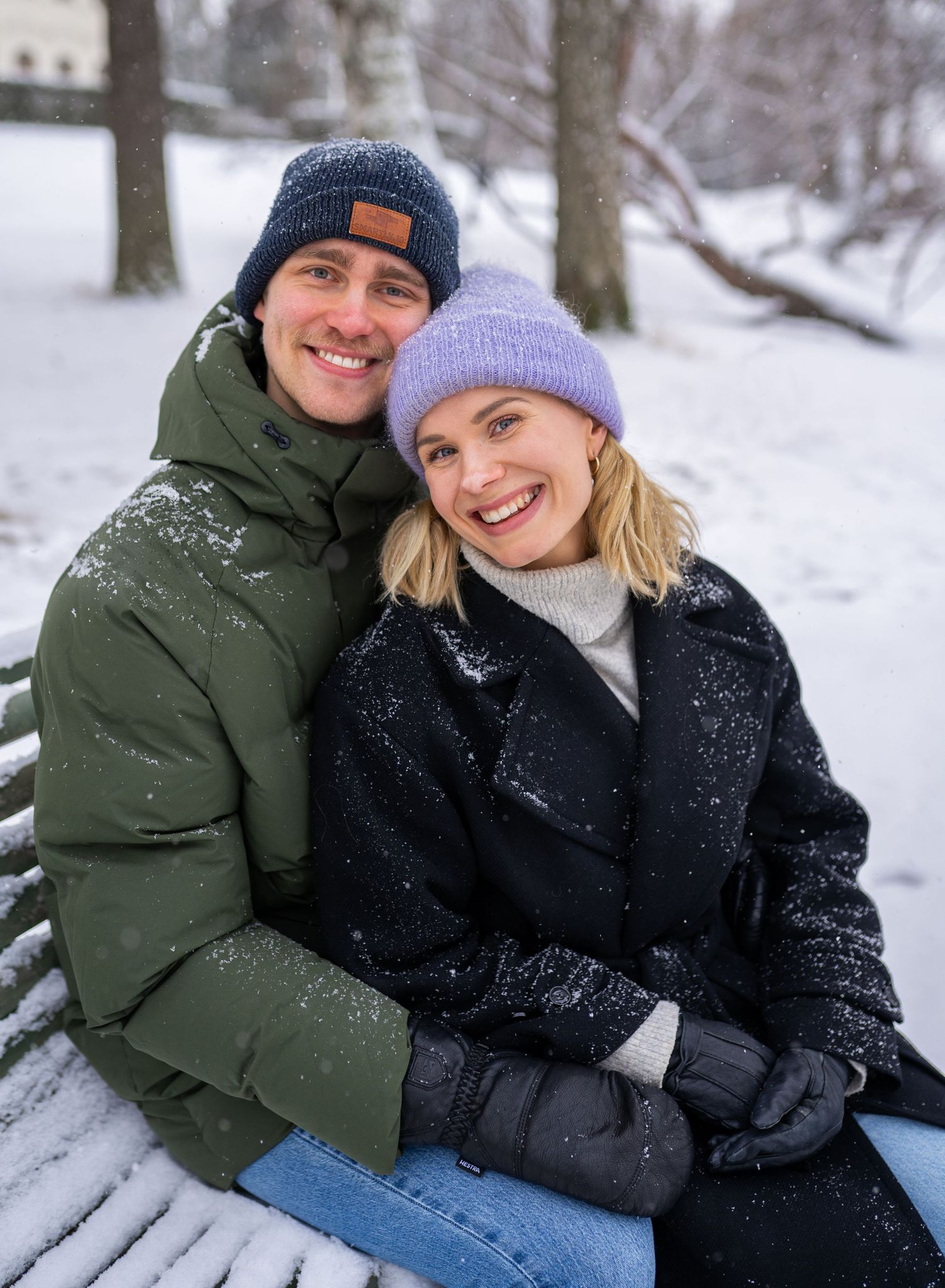 Happy couple sitting on snowy park bench wearing winter hats and puffer jackets, smiling at camera.