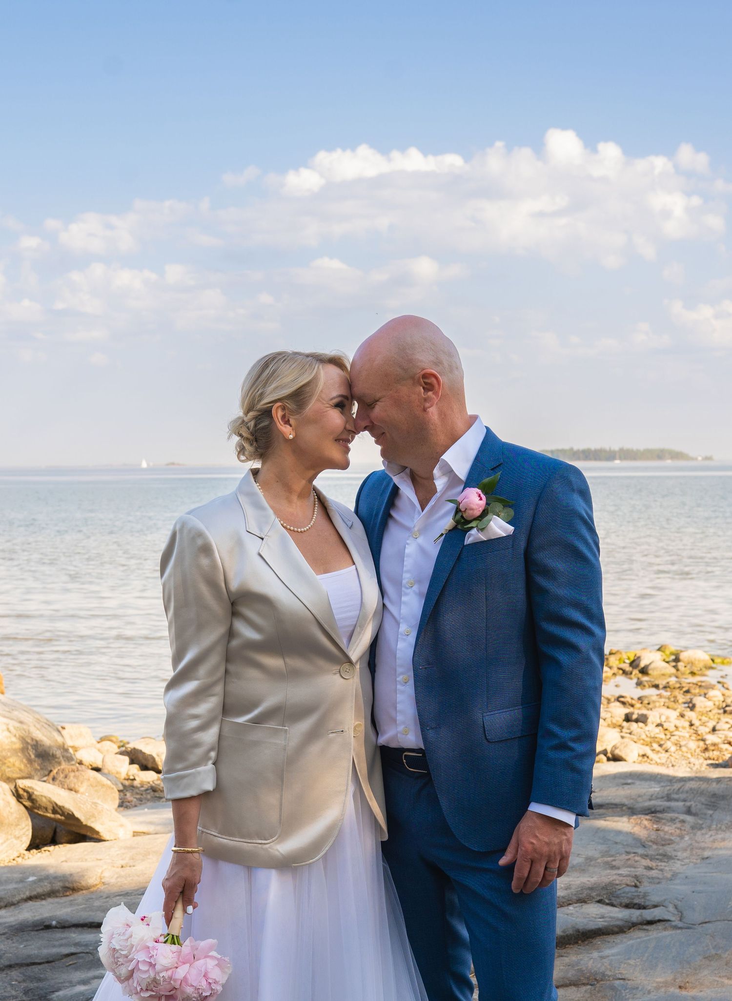 Bride and groom touch foreheads romantically by the rocky seaside, she in gold jacket, he in blue suit with boutonniere.