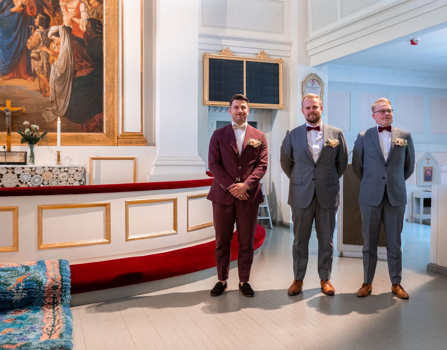 Groom in burgundy suit standing with two groomsmen in gray suits at church altar.