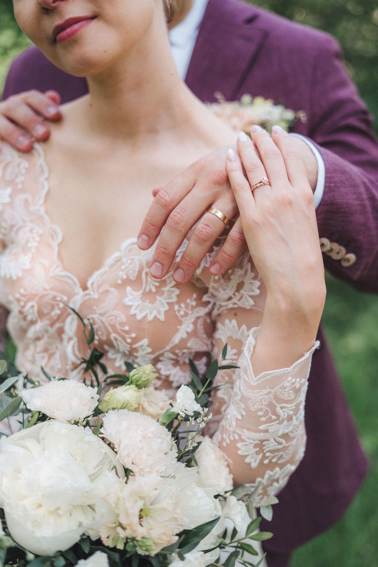 Close-up of bride and groom hands with wedding rings, bride in lace dress holding white peony bouquet.