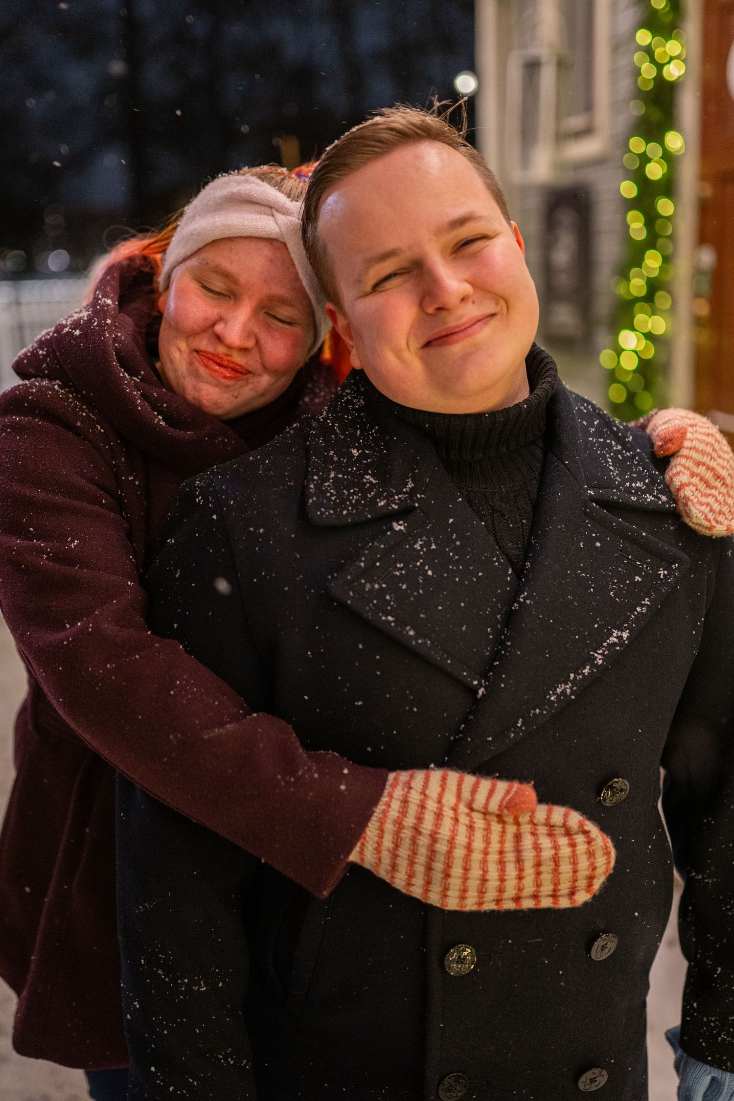 Two people hugging and smiling in the snow at night with Christmas lights in the background.
