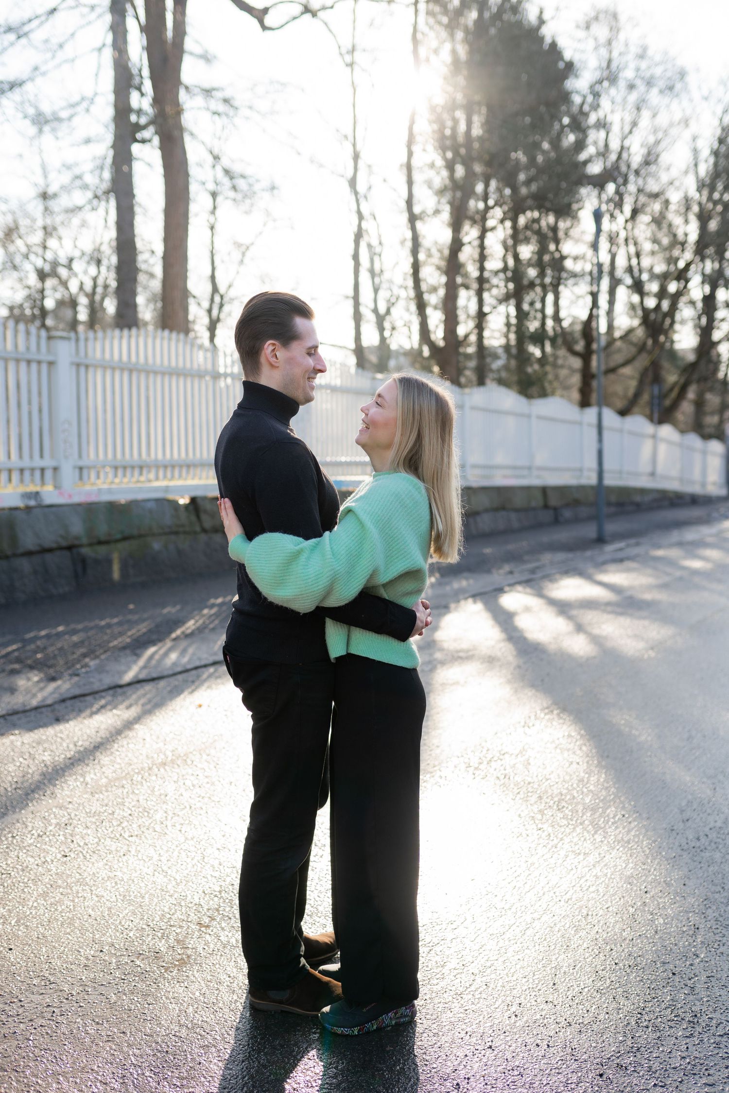 Couple embracing on a sunlit road, woman in mint green sweater and man in black, surrounded by trees and white fence.