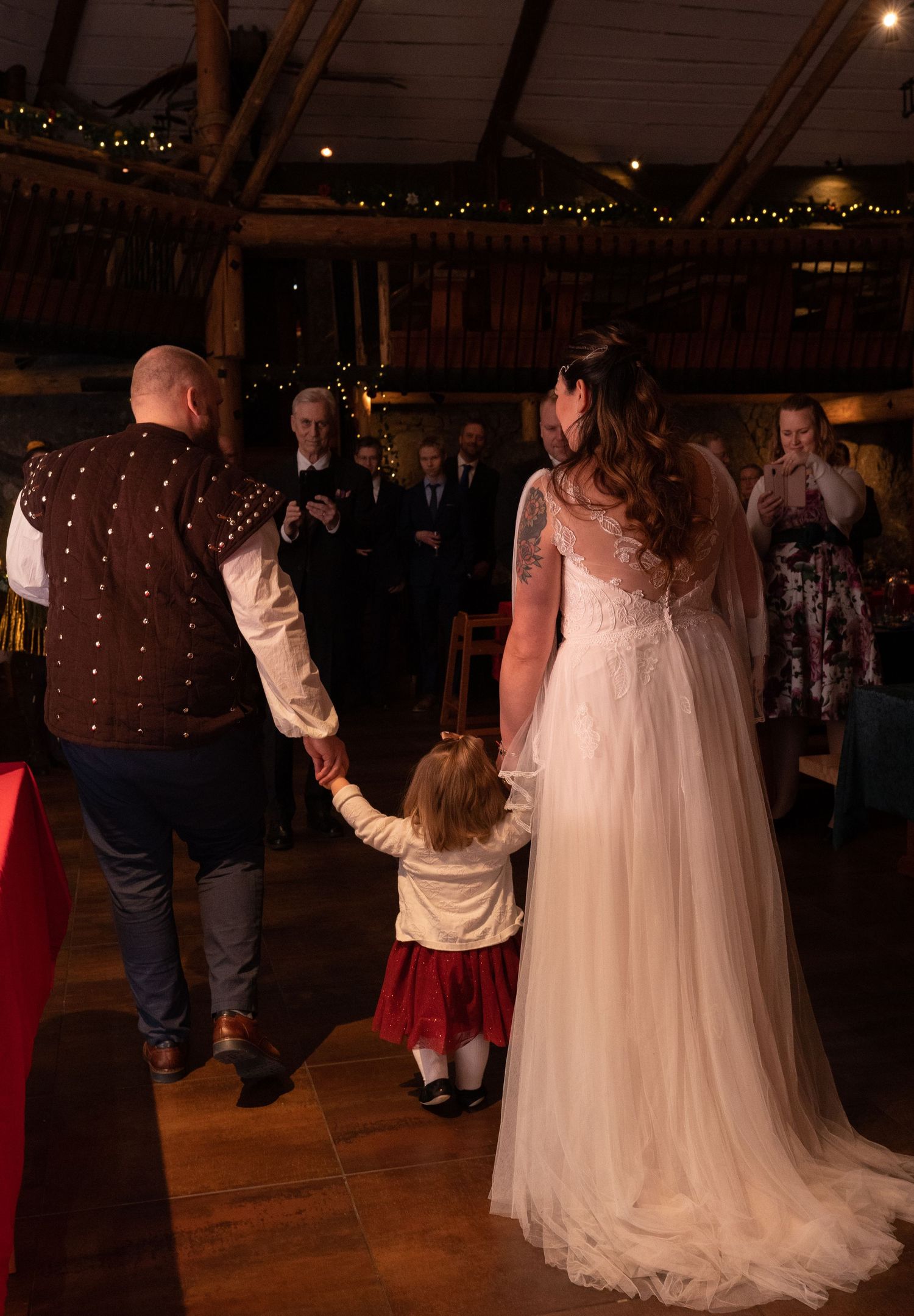 Bride and groom holding hands with young child at rustic barn wedding reception with fairy lights.