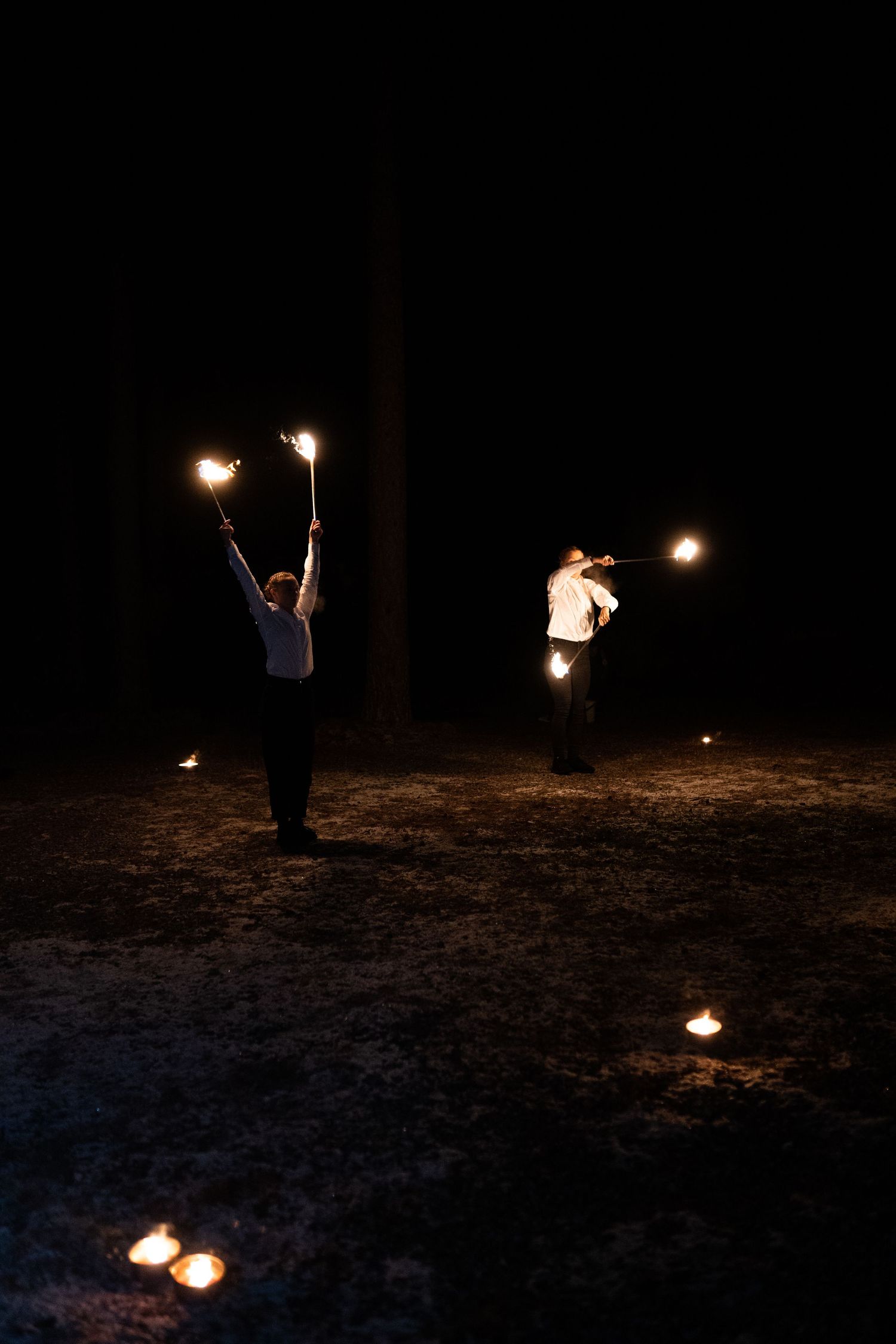 Two performers doing a fire show at night, holding burning torches with candles on the ground around them.