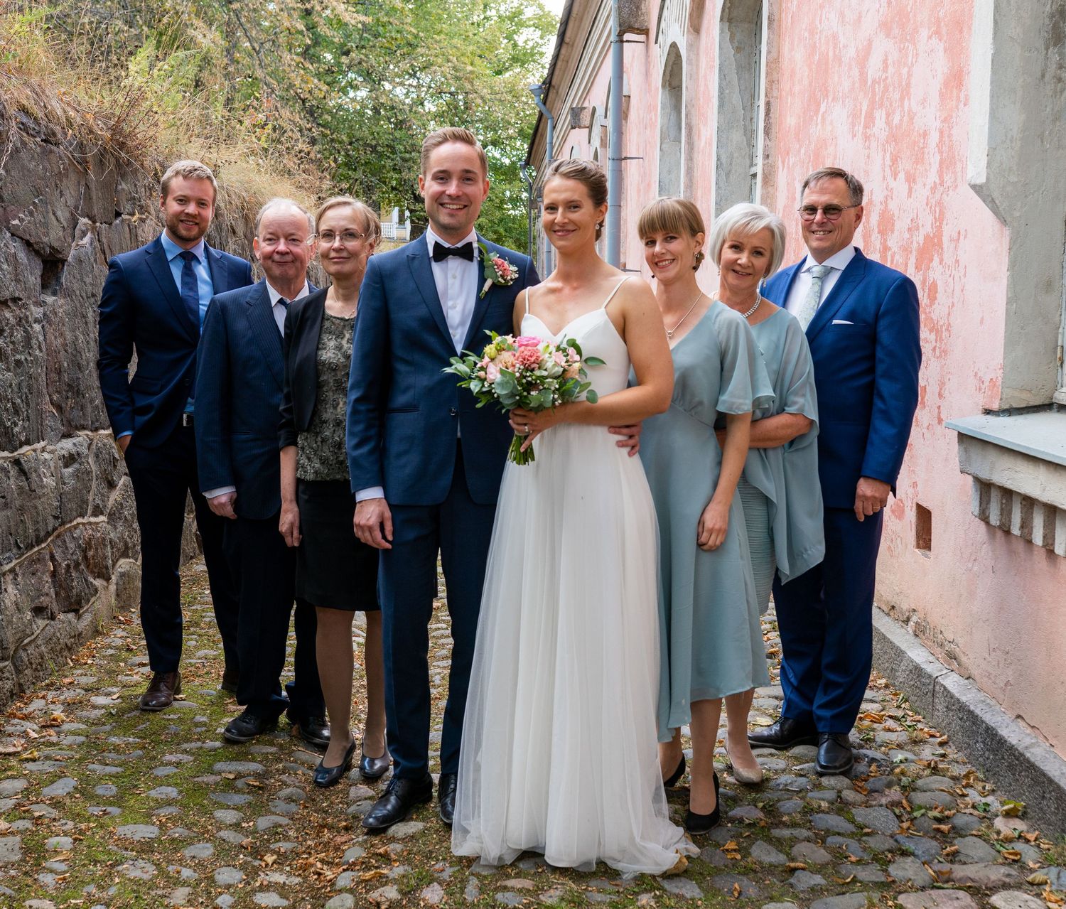 Wedding party of seven poses together outdoors by pink building with bride in white and groom in navy.