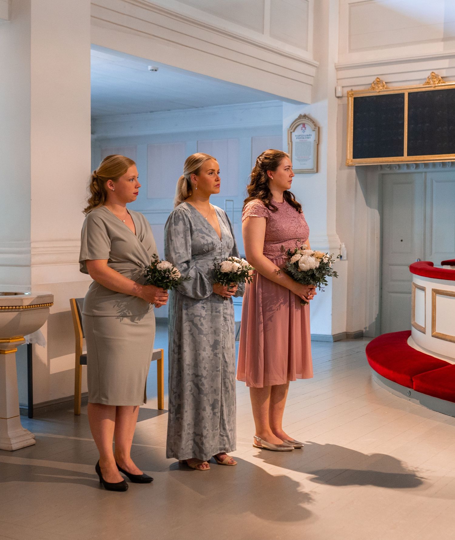Three bridesmaids in pastel dresses holding white floral bouquets stand inside elegant white church interior.