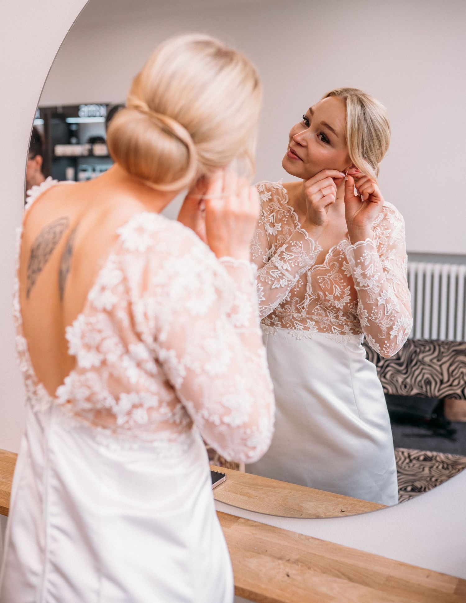 Blonde bride in lace wedding gown putting on earrings while looking in a round mirror.