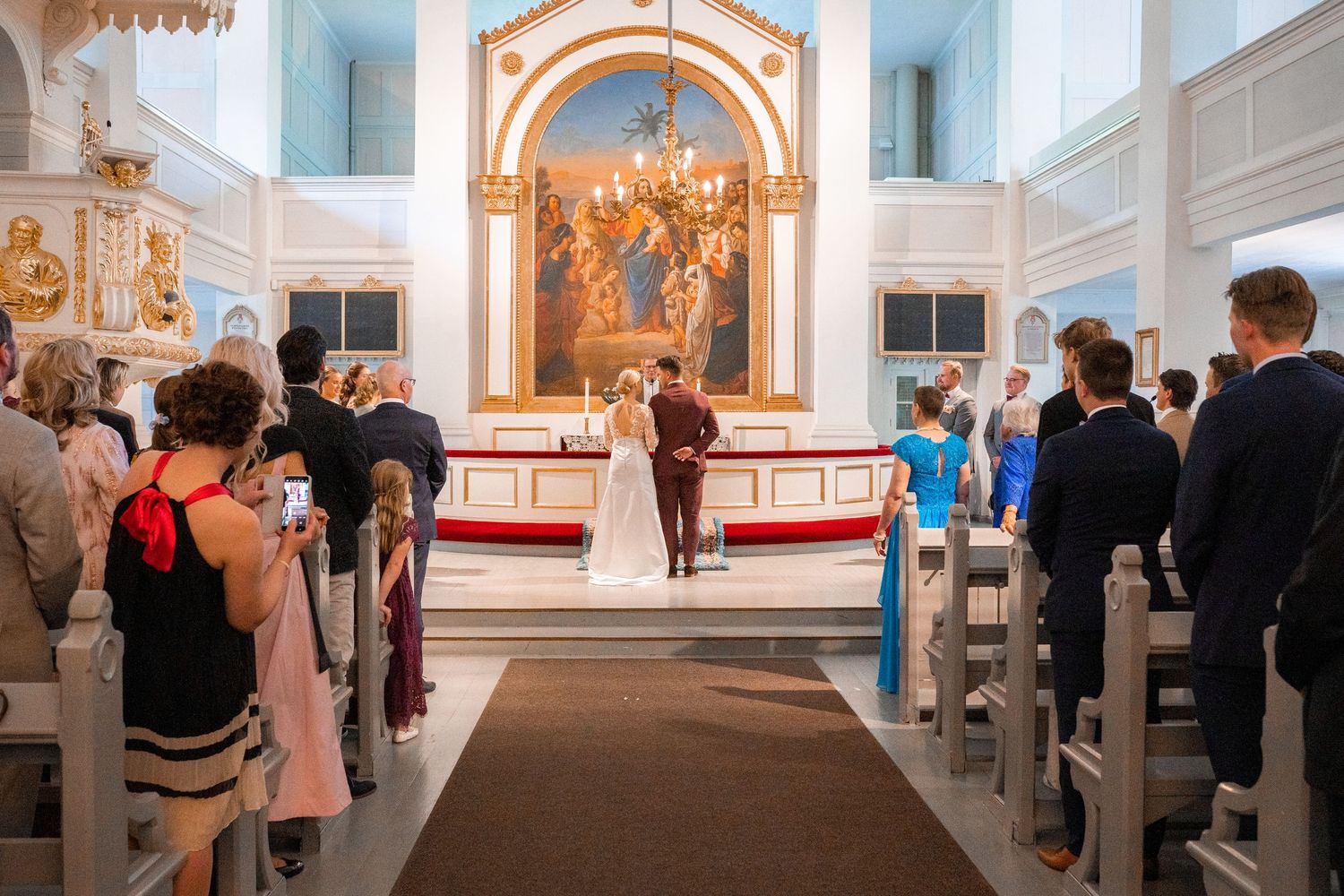 Wedding ceremony in a beautiful white church with ornate altar, guests seated in pews watching the couple exchange vows.