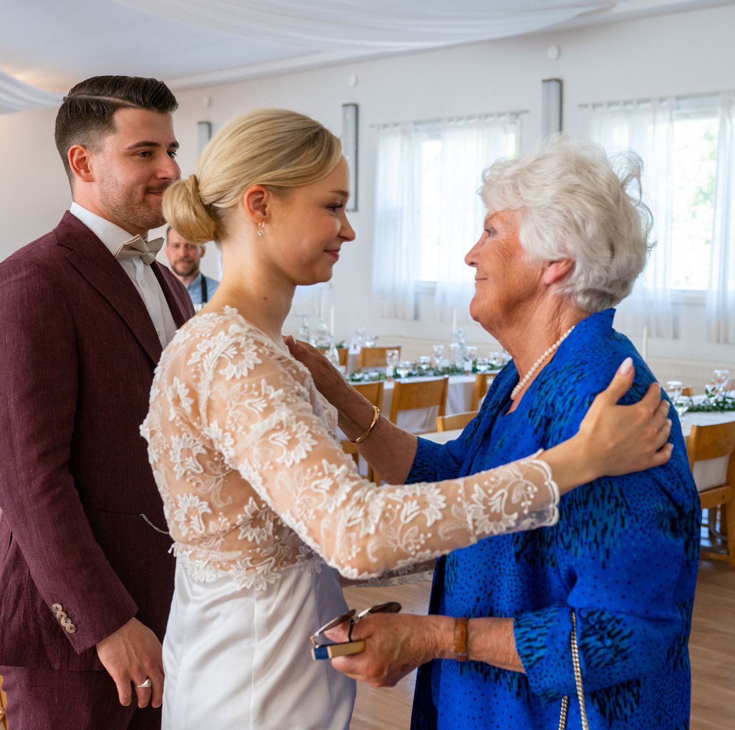 Bride in lace gown warmly greets elderly woman in blue at wedding reception while groom watches.