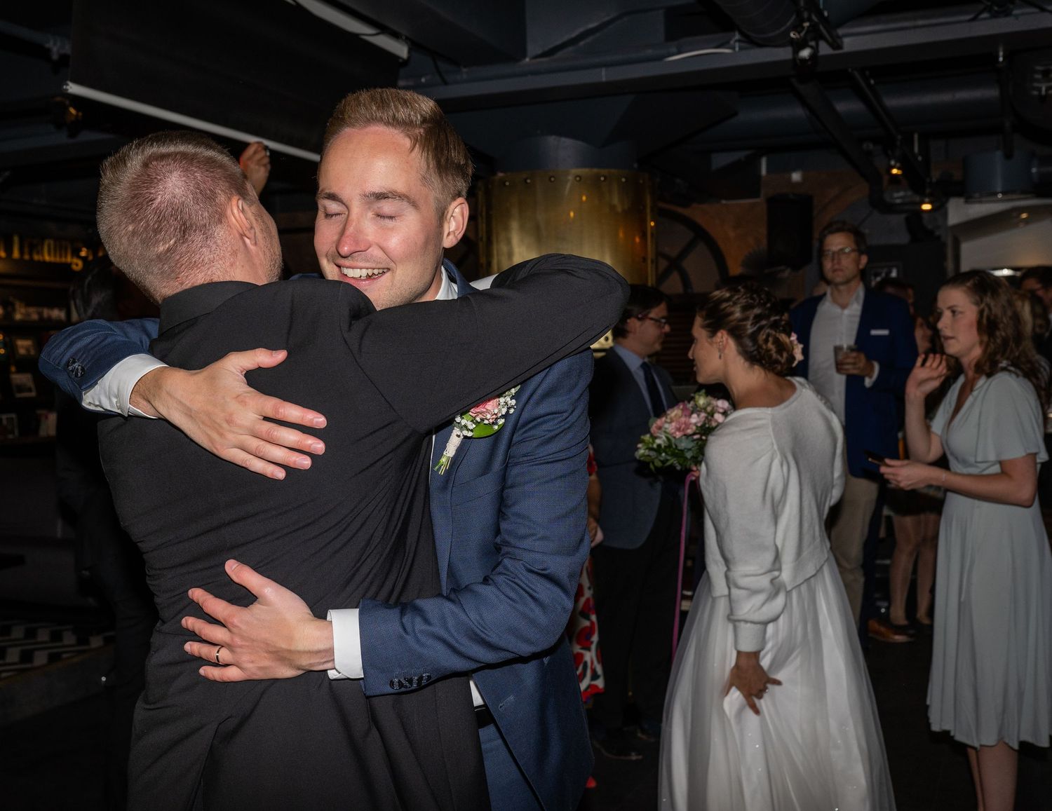 Groom in navy suit shares warm embrace with guest while bride stands nearby at wedding reception.