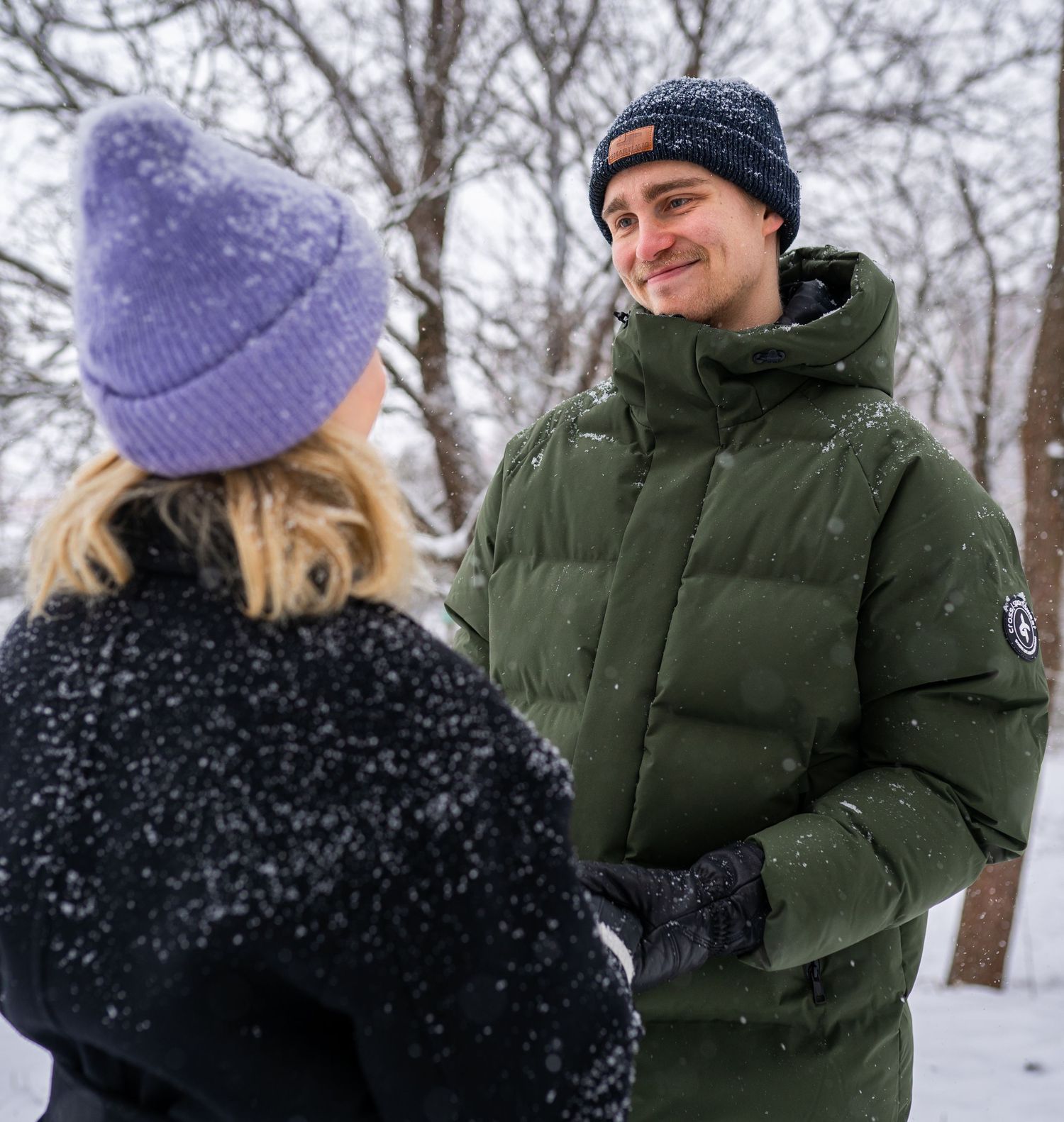 Couple smiling at each other in snowy winter park, wearing warm hats and jackets.