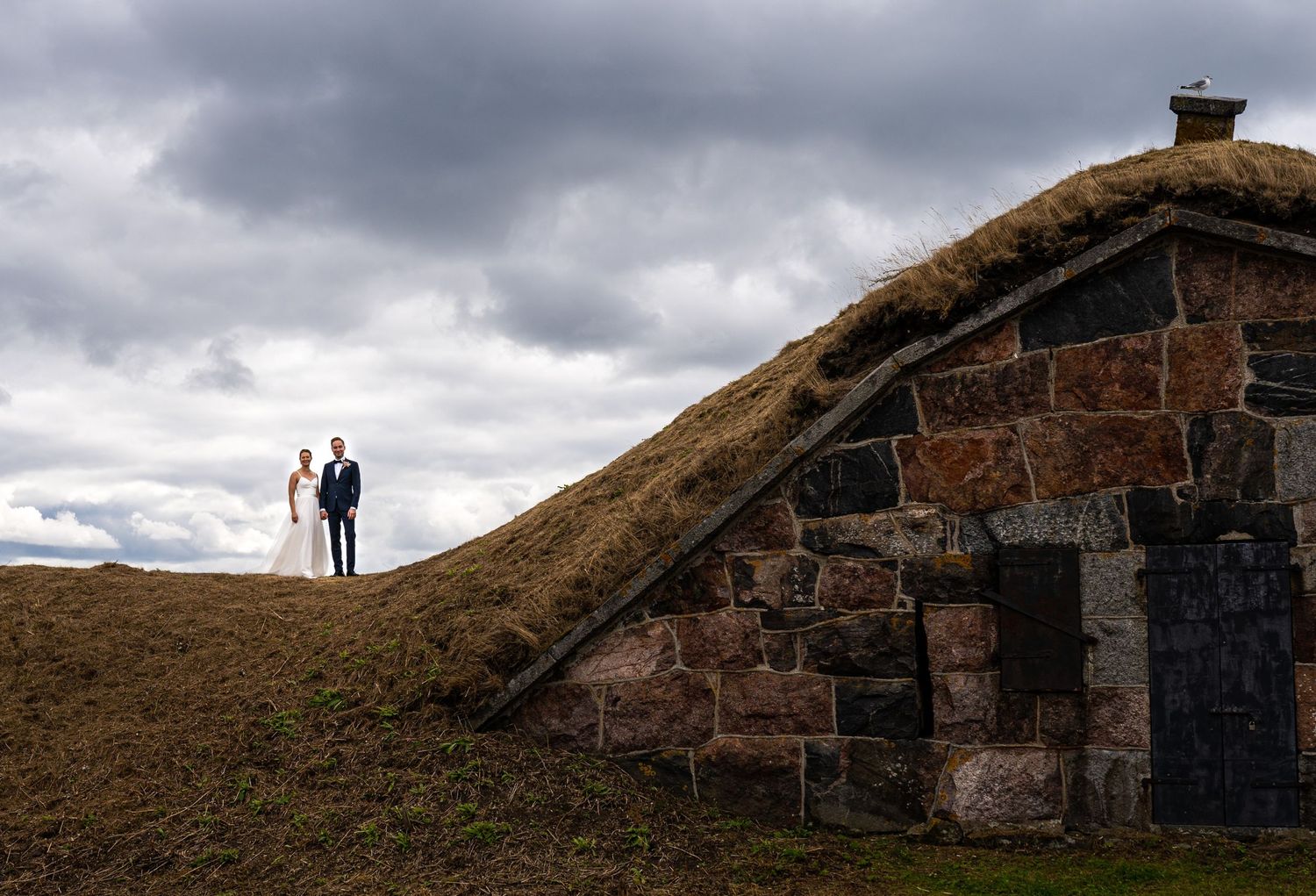 Bride and groom stand atop a rustic stone building with grass roof under dramatic cloudy skies in Iceland.
