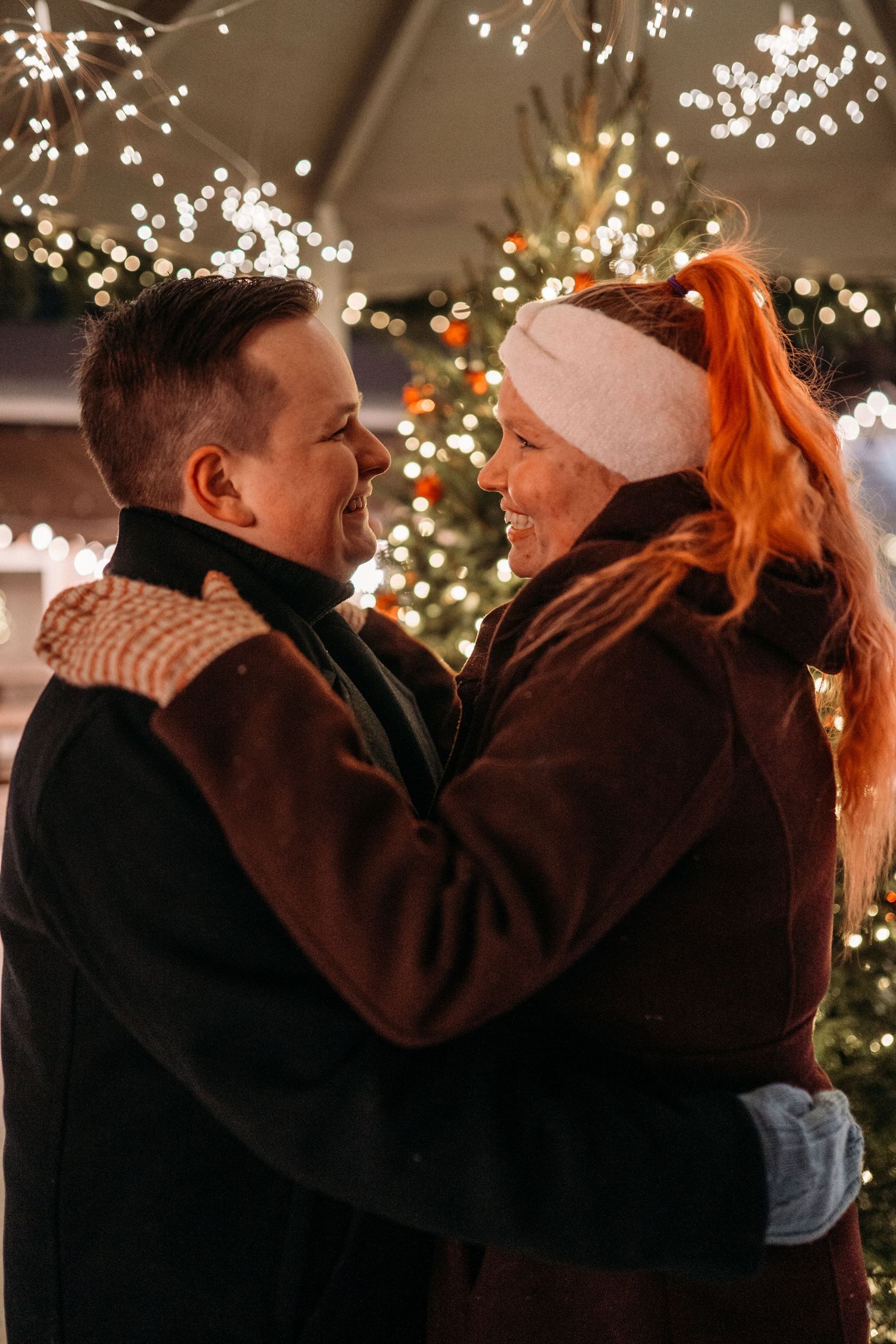 Couple embracing and smiling at each other in front of Christmas tree lights at night.