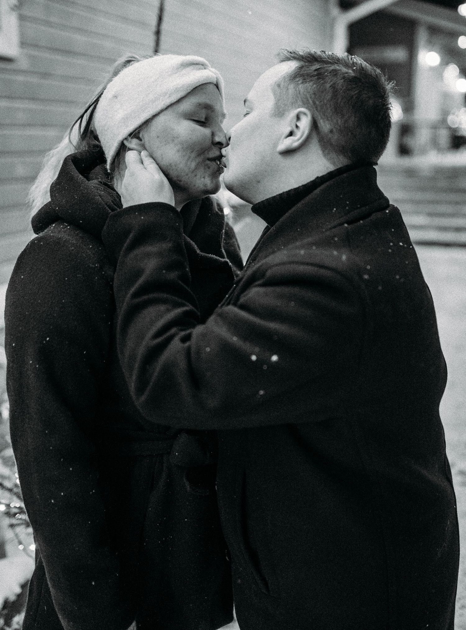 Black and white photo of a couple sharing a romantic kiss outdoors in winter, woman wearing a white hat.
