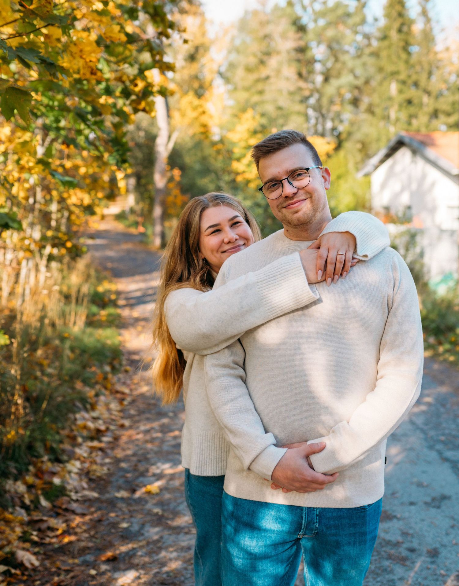 A couple in matching cream sweaters embraces warmly on a tree-lined path surrounded by golden autumn foliage.