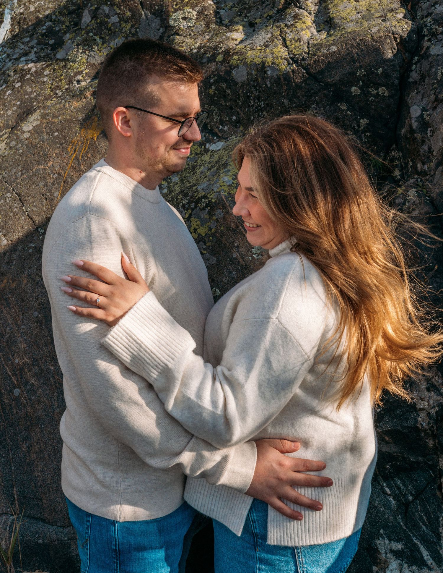 A couple in matching cream sweaters and jeans embrace and smile at each other against a rocky outdoor backdrop.