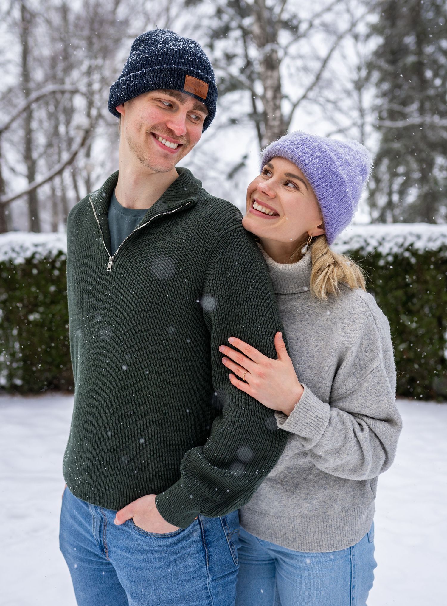 Smiling couple in winter beanies embracing outdoors in snowy weather, wearing cozy sweaters.