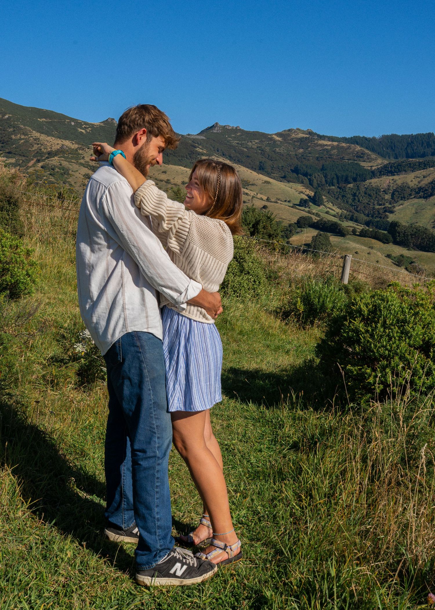 Romantic couple embraces on lush green hillside with rolling mountain landscape behind them.